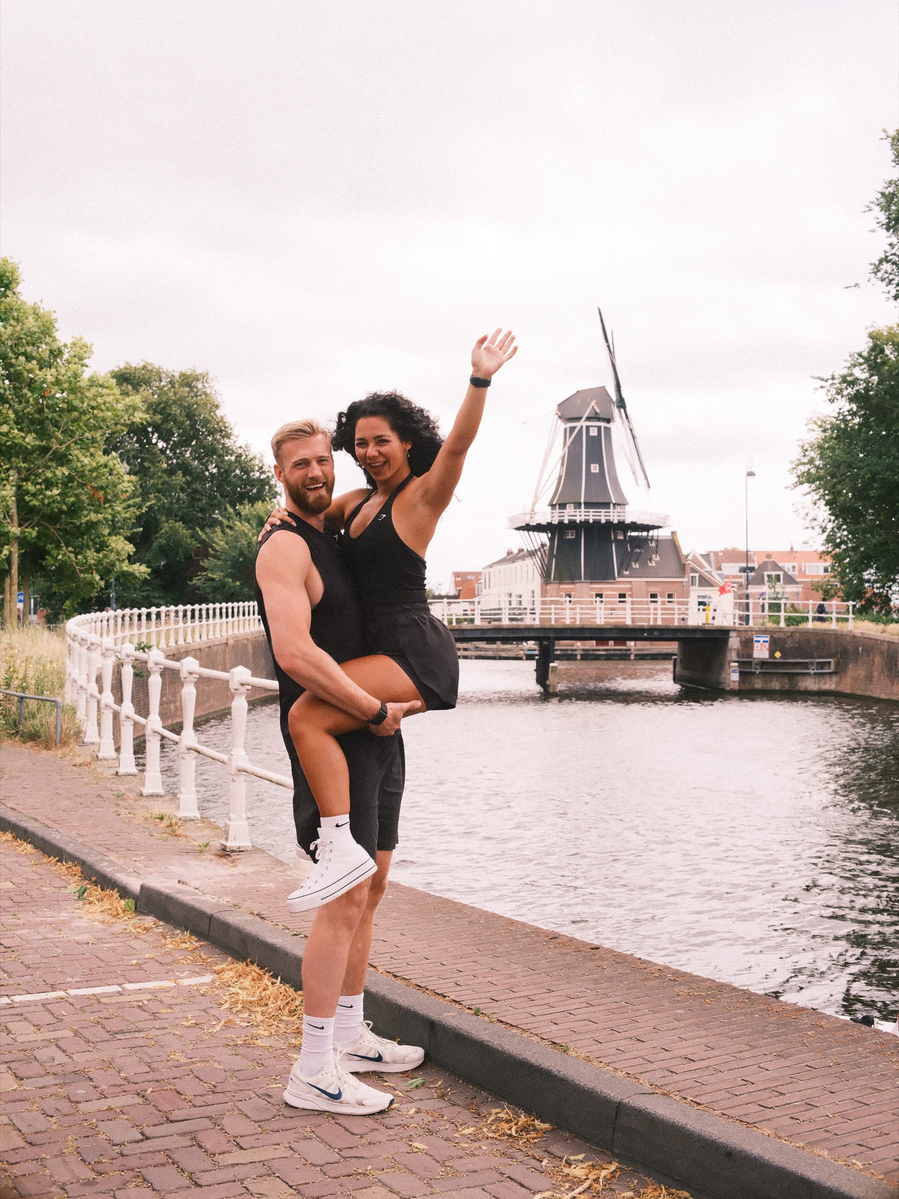 Man tilt zijn vriendin op bij een wit hek terwijl zij een arm in de lucht steekt, met de molen aan de rivier erachter