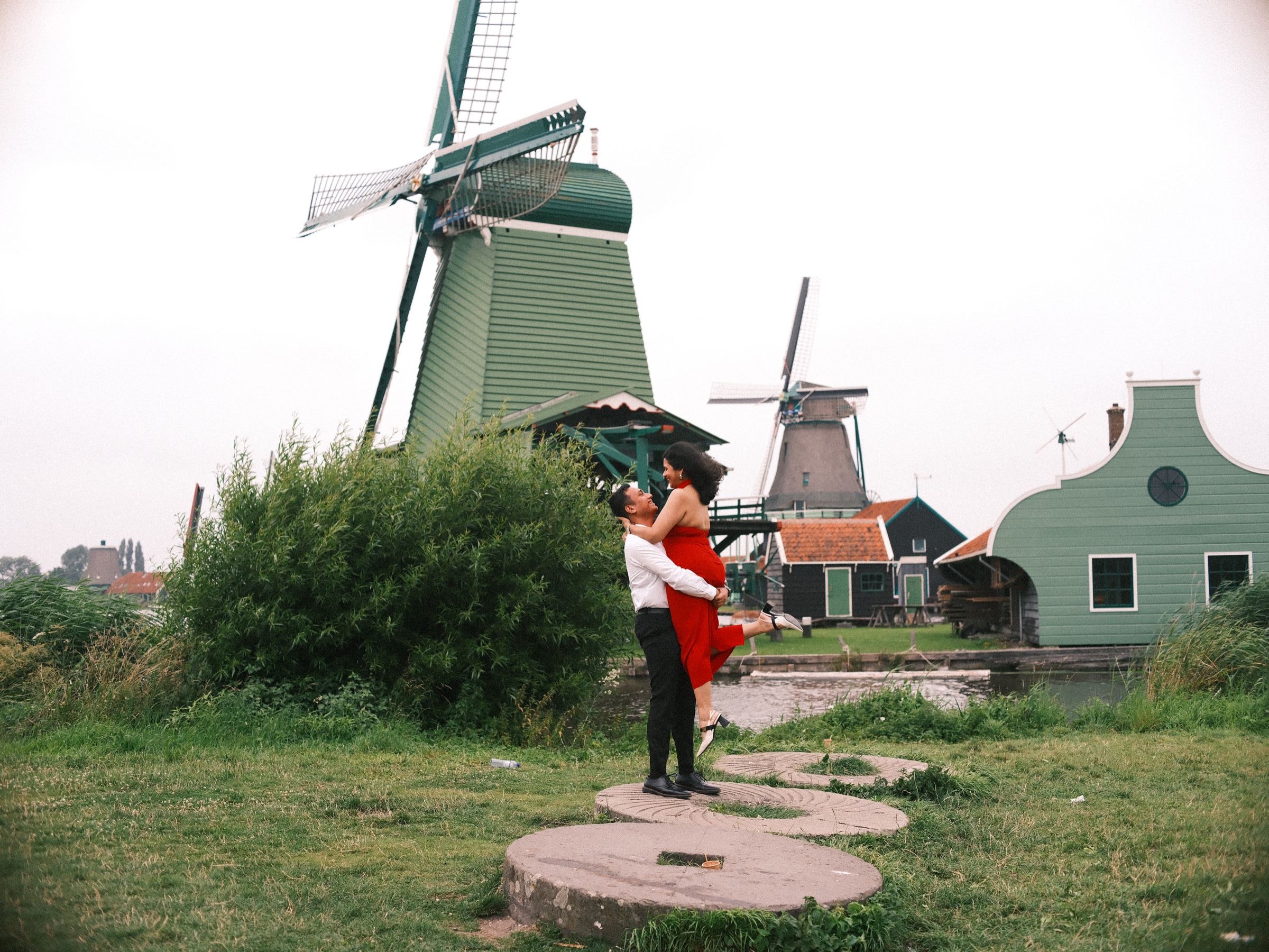 Man lifting his partner in a red dress on old millstones in the grass, the big green windmill of the Zaanse Schans behind them