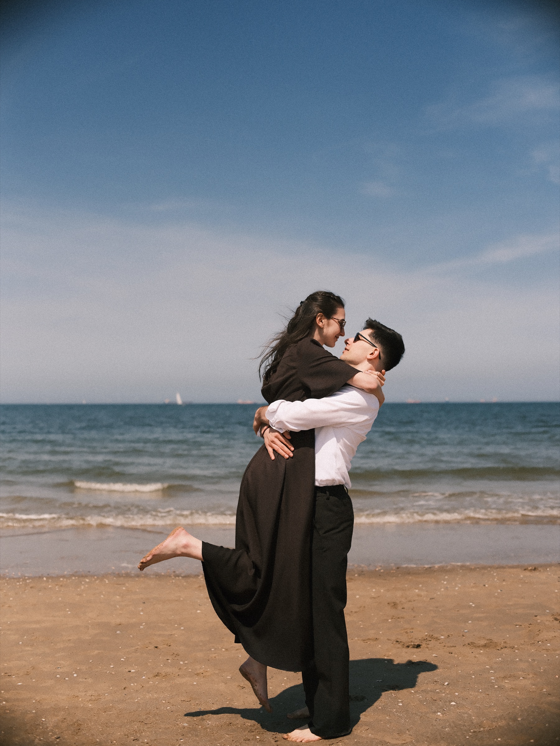 Man lifting his fiancée off the sand as she kicks one bare foot into the air, the last frame of their beach proposal