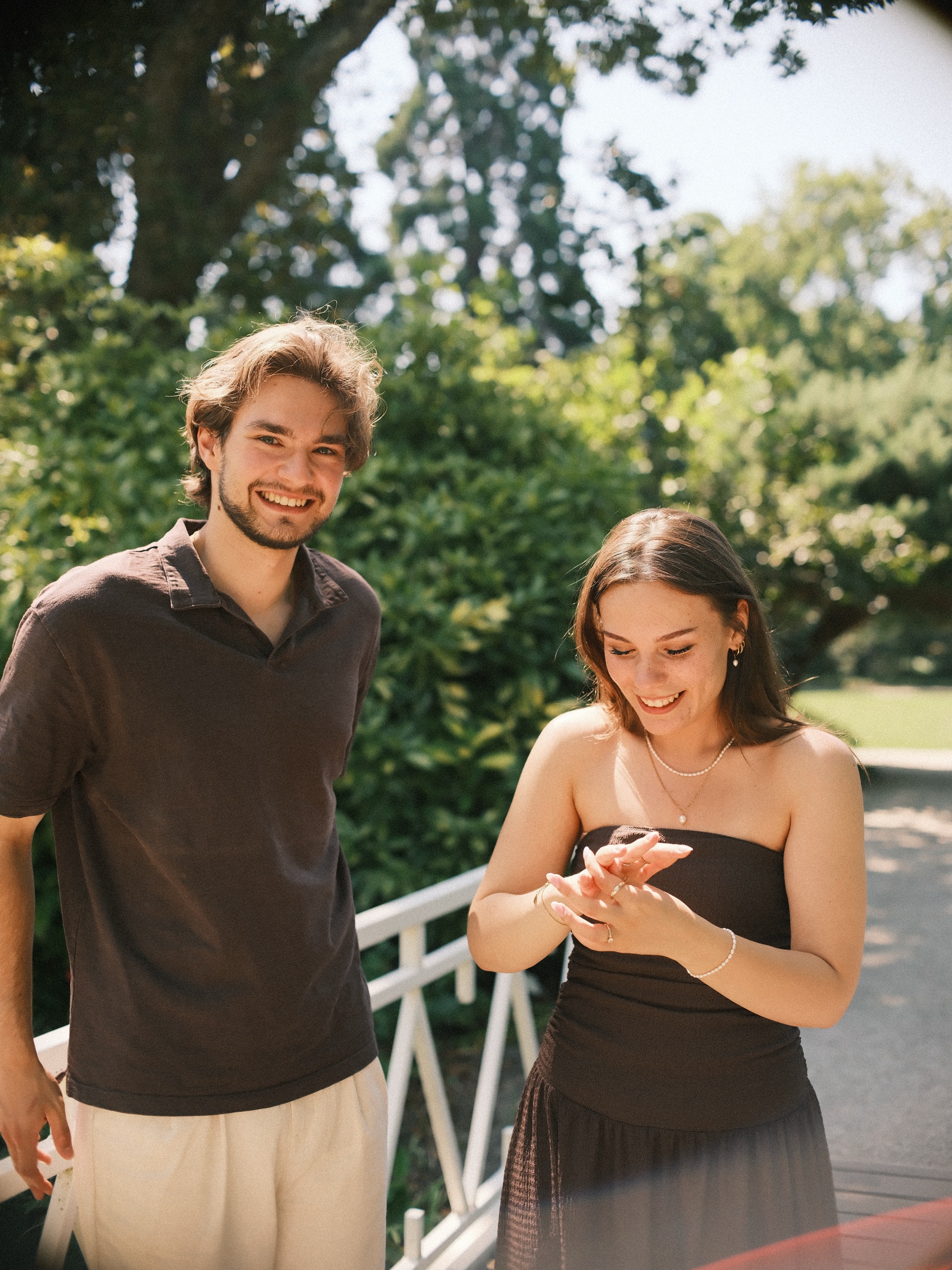 She looks down at the new ring on her hand while he smiles at the lens, the white railing behind them just after the surprise proposal