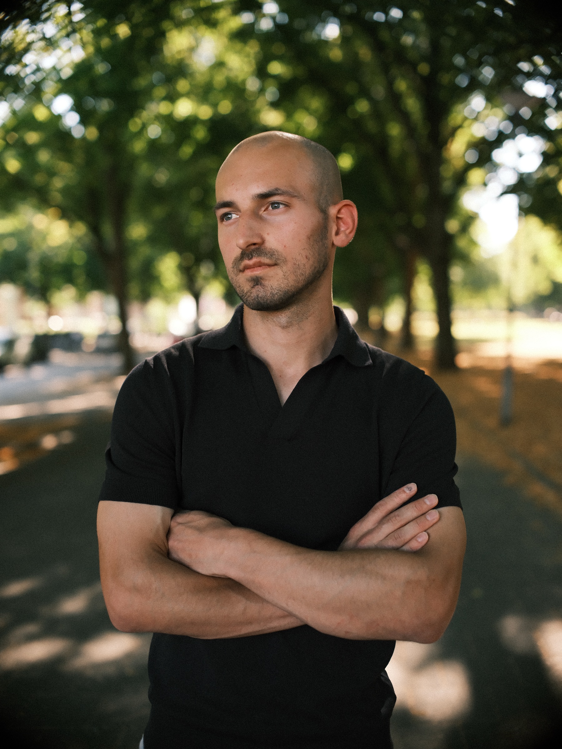 Man in een zwarte polo met de armen over elkaar onder oude bomen in Rotterdam, met lage avondzon op het pad, tijdens een solo fotoshoot