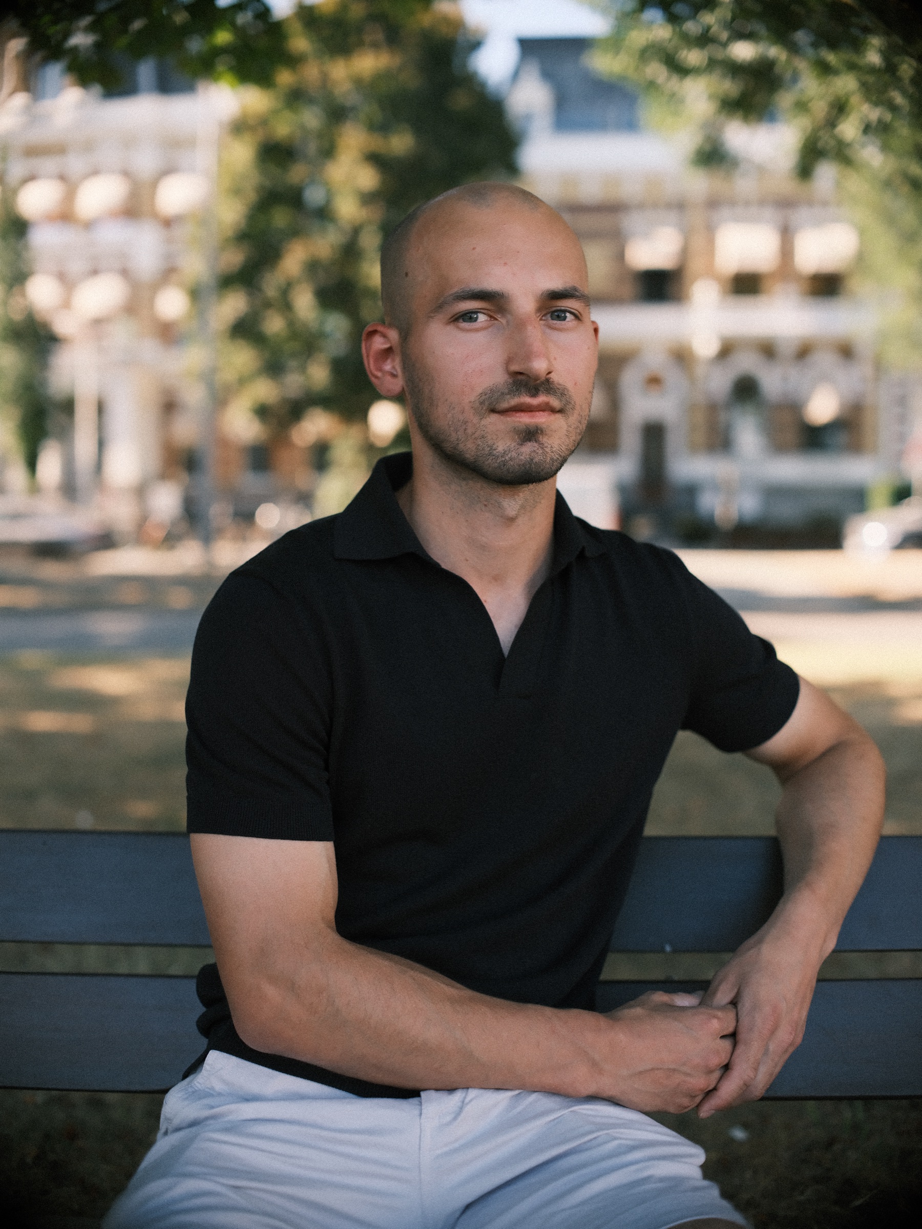 Man in a black polo shirt on a bench under trees, a grand old building softly blurred behind him, a portrait photo session in Rotterdam