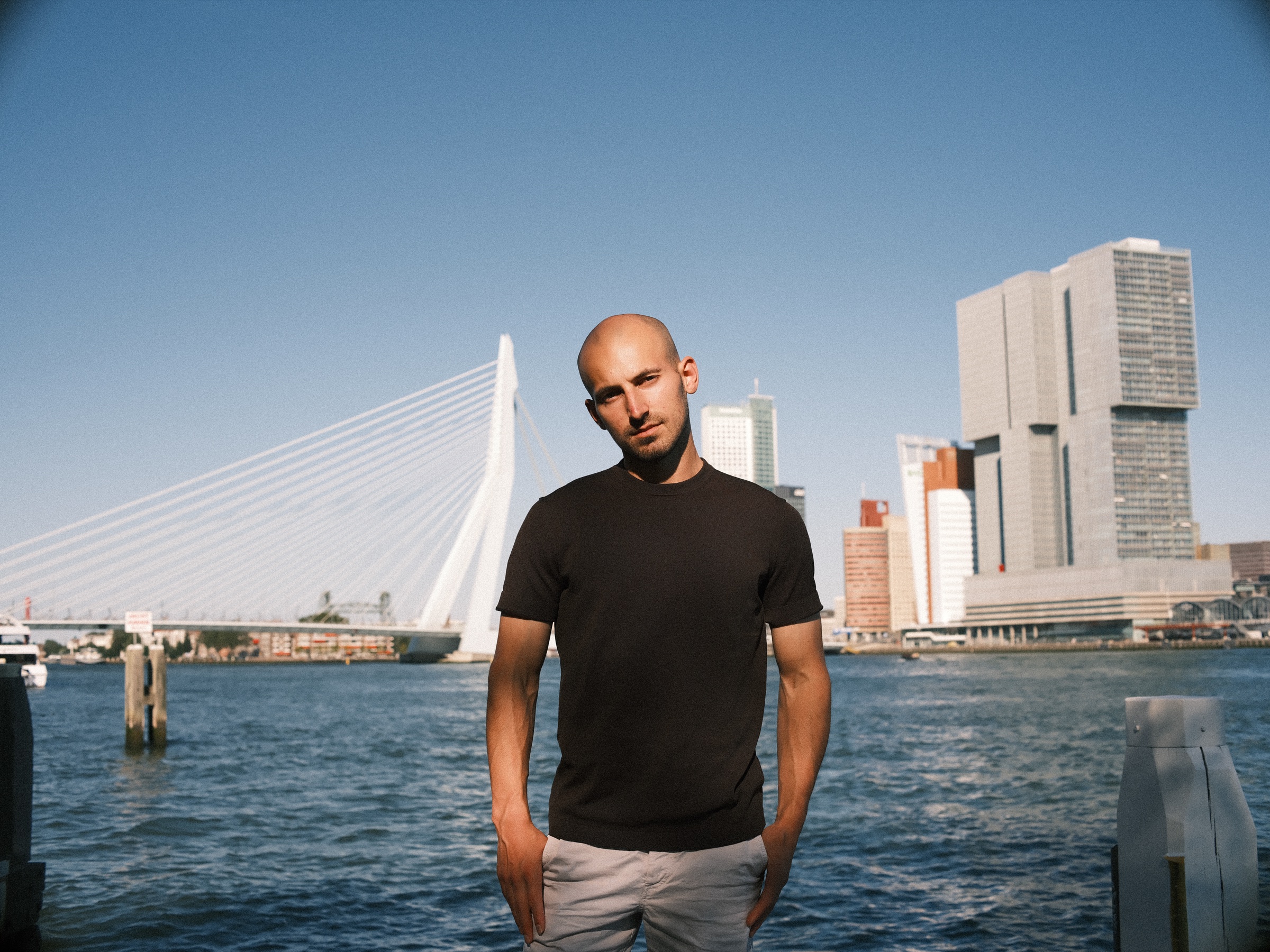 Man in een zwart T-shirt met zijn handen in de zij aan de Maas, met de Erasmusbrug en De Rotterdam achter hem