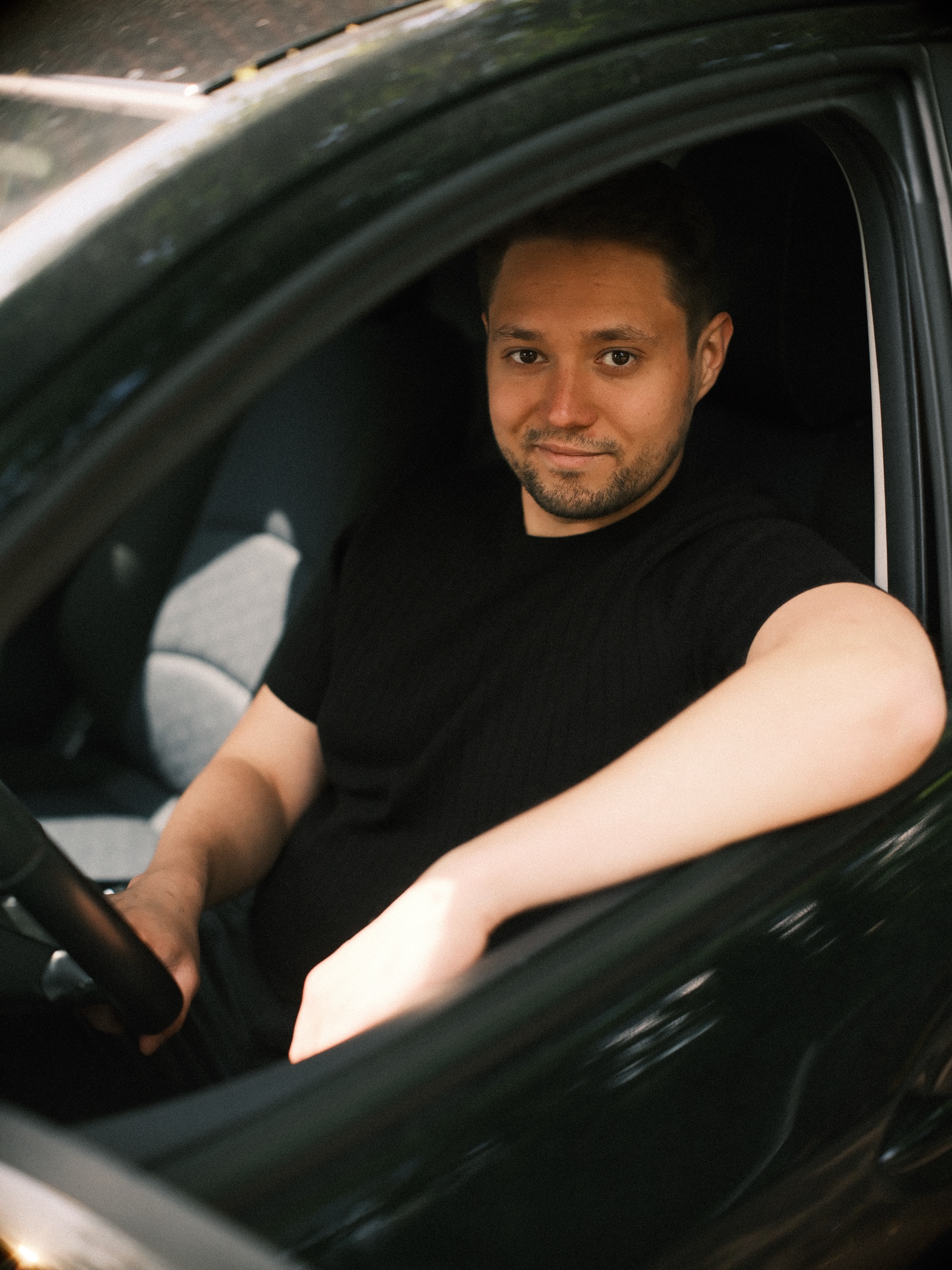 Man in zwart T-shirt lacht vanaf de bestuurdersstoel van zijn auto, met een arm in het open raam, tijdens zijn fotoshoot in Haarlem