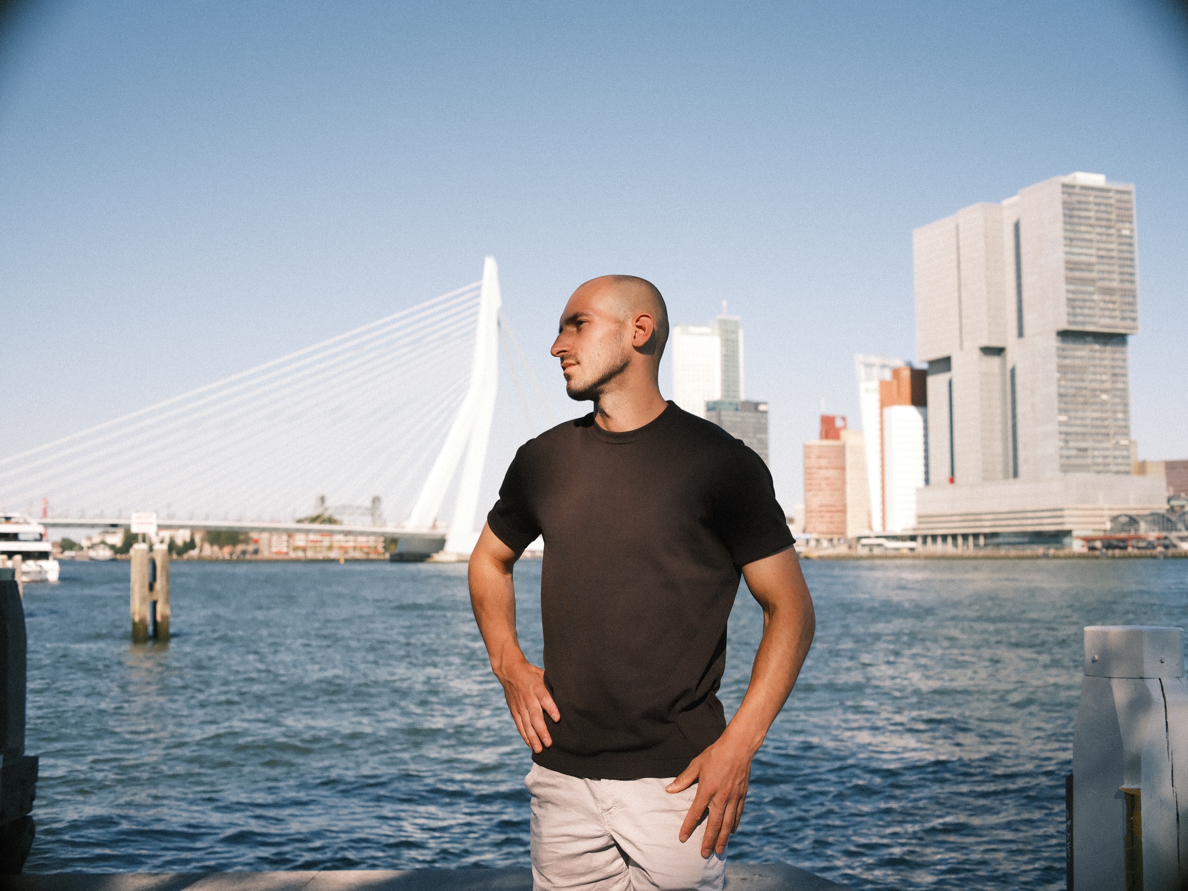 Man in een zwart T-shirt staat met de handen in de zij aan de Maas, met de Erasmusbrug en de torens van De Rotterdam achter hem op een avond in augustus