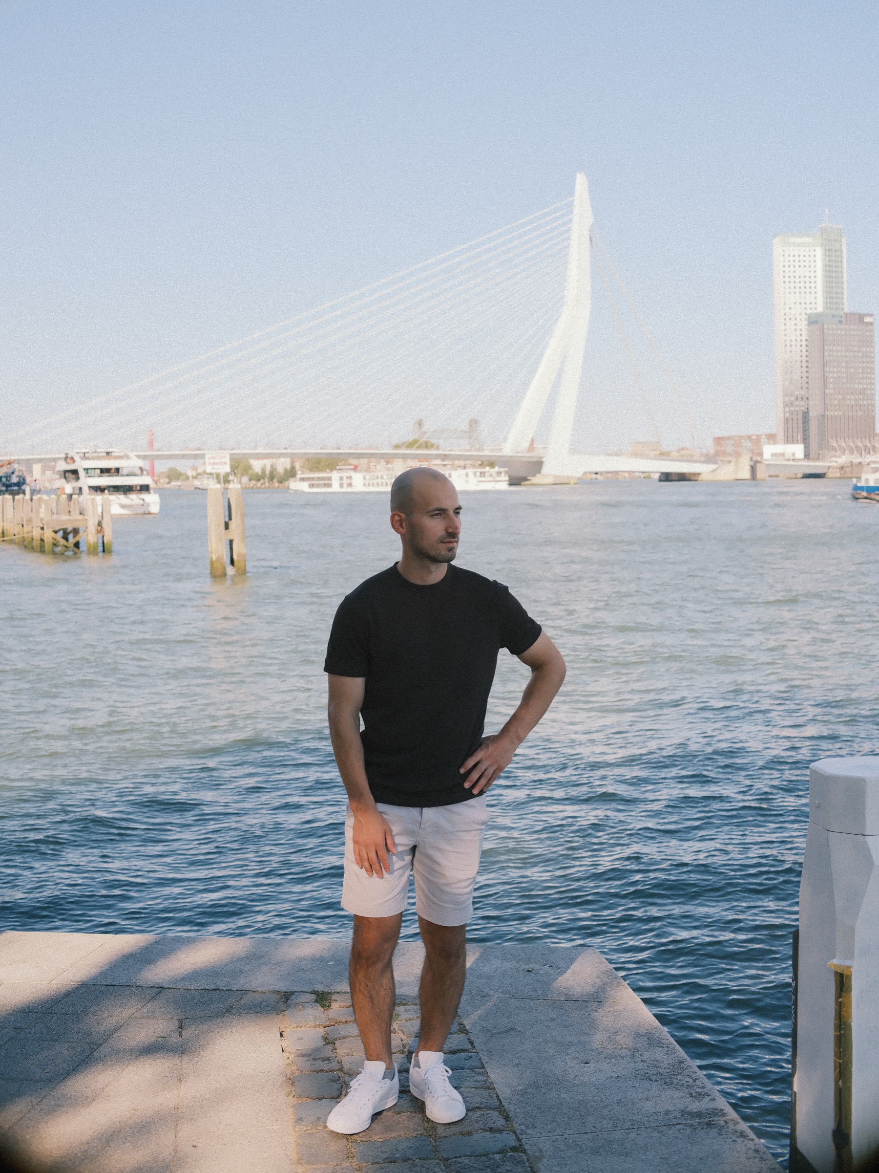 Staand op een stenen kade aan de Maas met zijn handen in de zij, met de Erasmusbrug achter hem op een heldere zomeravond