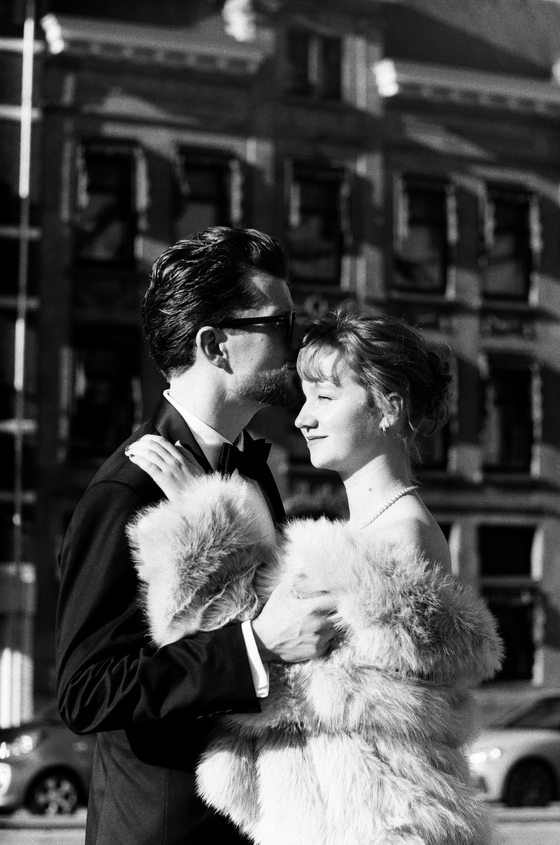 He kisses her temple as she smiles with her eyes half closed, pearls and a fur coat, an old facade behind them on Ilford film