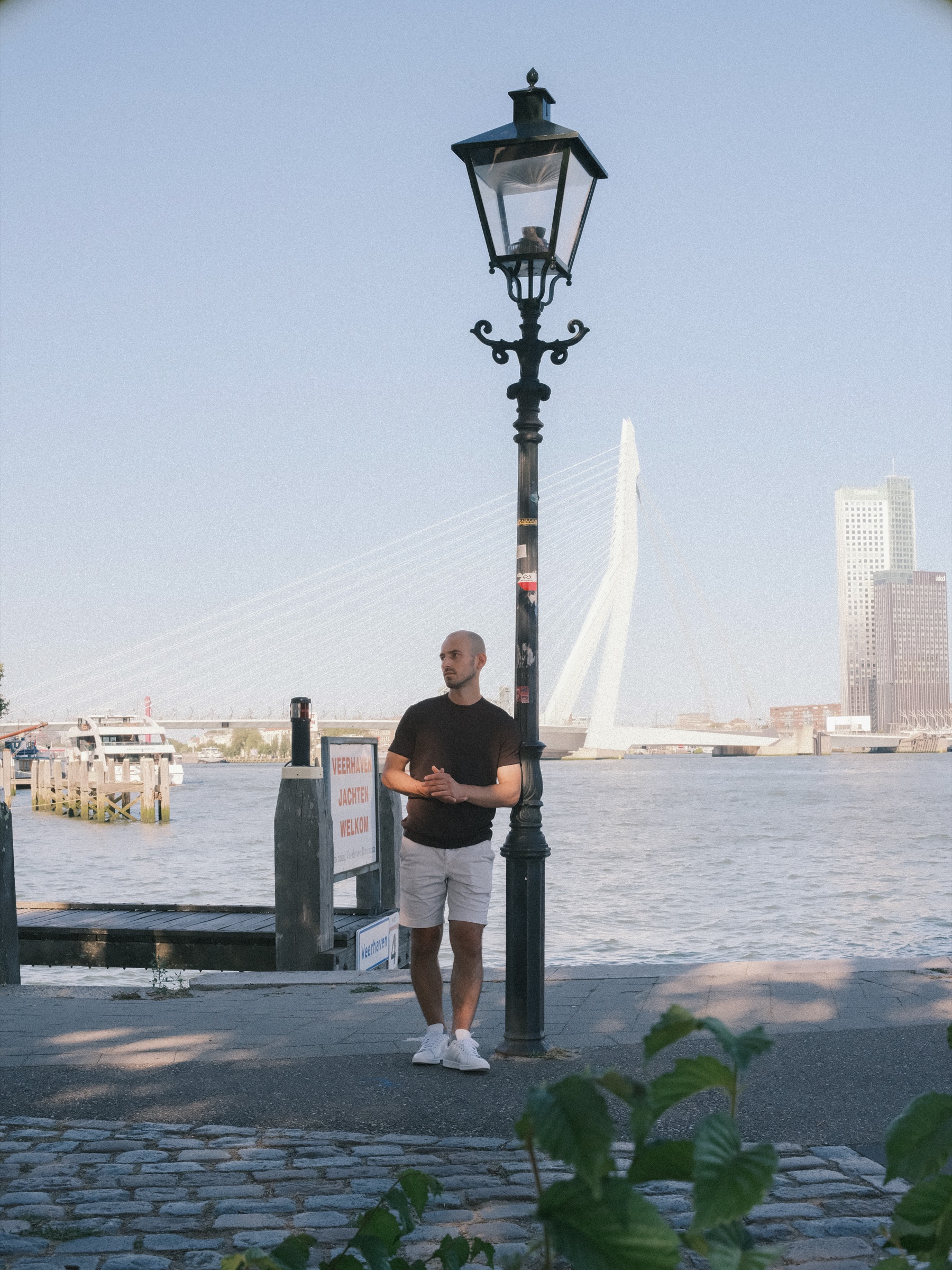 Man by an old lamppost and a Veerhaven welcome sign on the cobbled Maas quay, the Erasmusbrug behind him in clear evening light