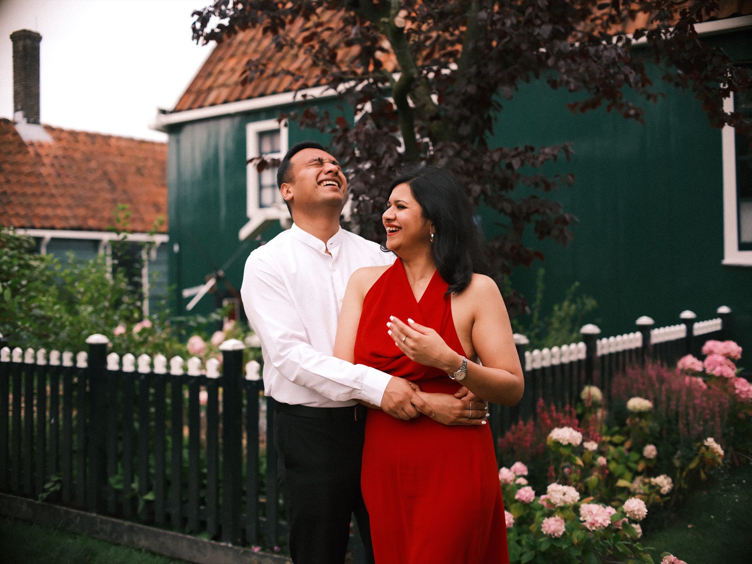 Man laughing with his head thrown back while she smiles up at him, a dark picket fence and pink hydrangeas around them