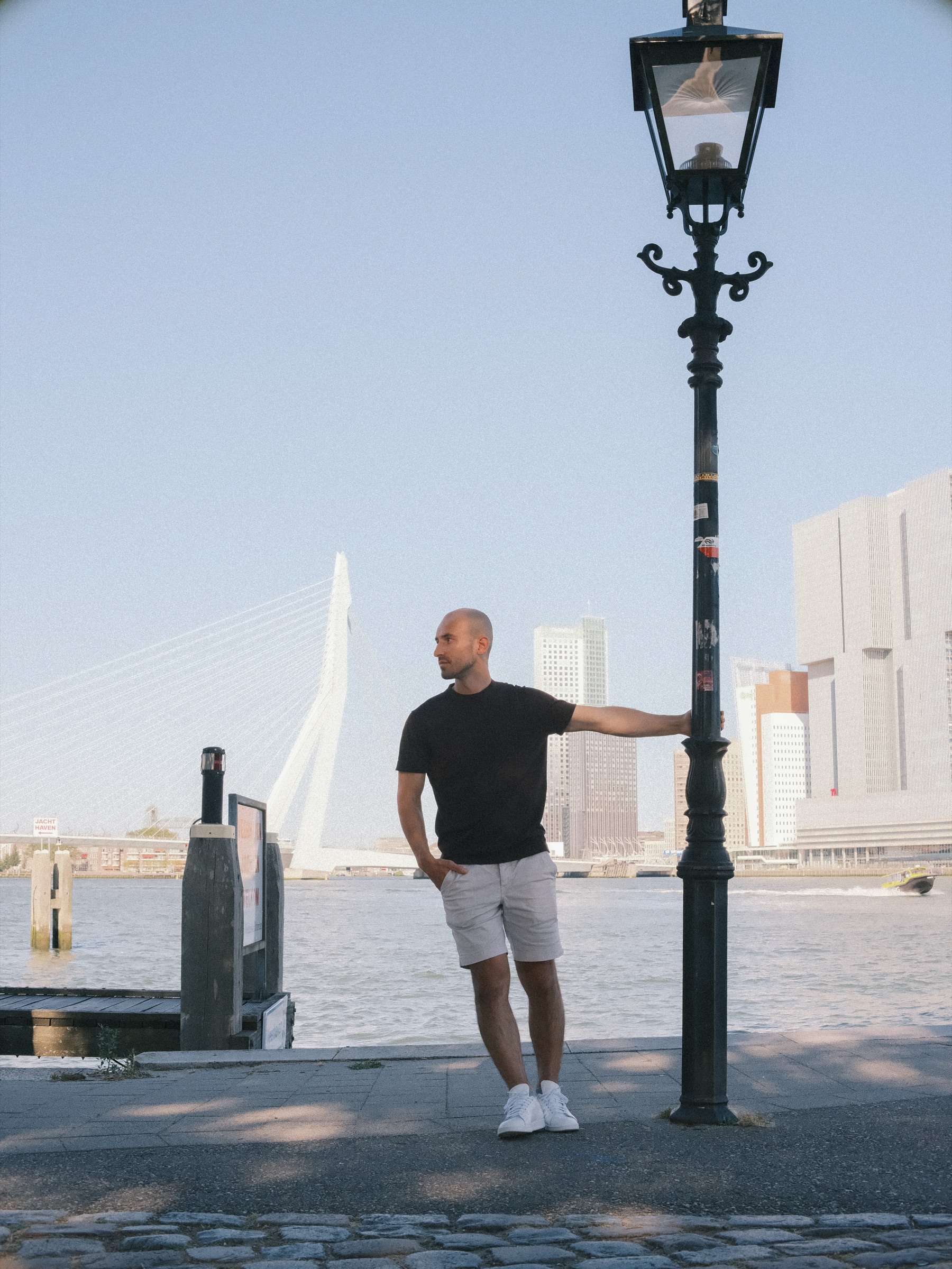 Eén hand op een oude zwarte lantaarnpaal op de kade, hij kijkt over de rivier, met de Erasmusbrug en De Rotterdam erachter