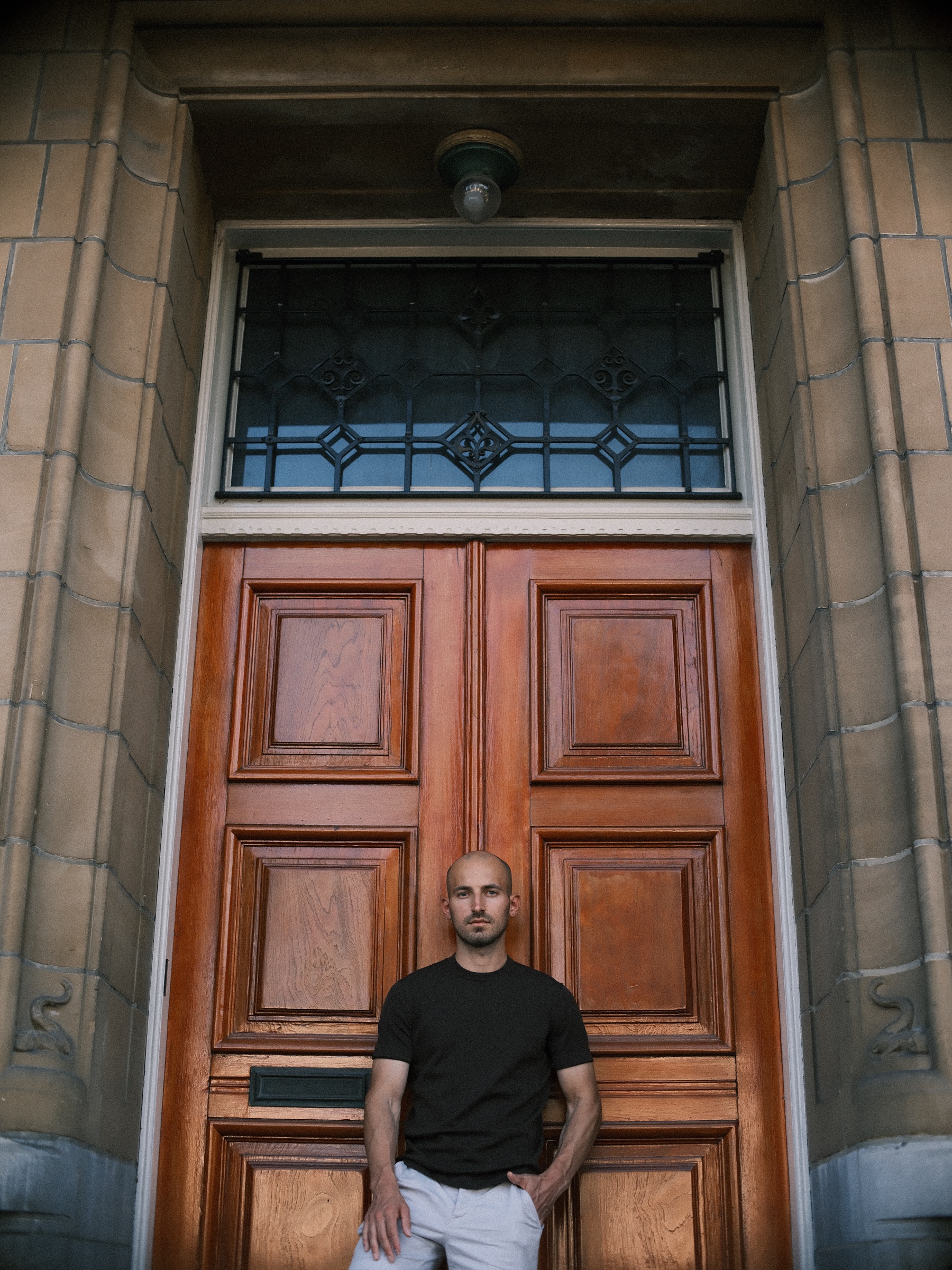 Man in a black T-shirt leaning on a tall red-brown wooden door, stained glass and an old lamp above him in a deep stone doorway
