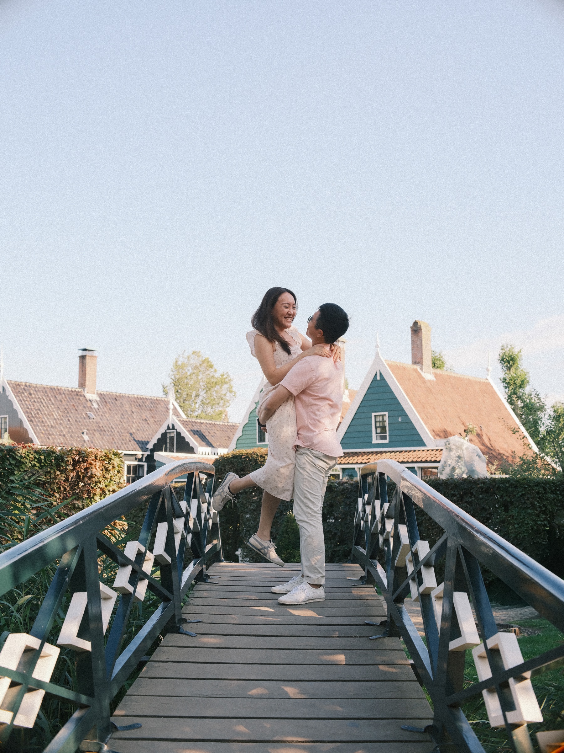 Man lifting his laughing fiancée on a wooden bridge with gray and white railings, the gabled roofs of the Zaanse Schans behind