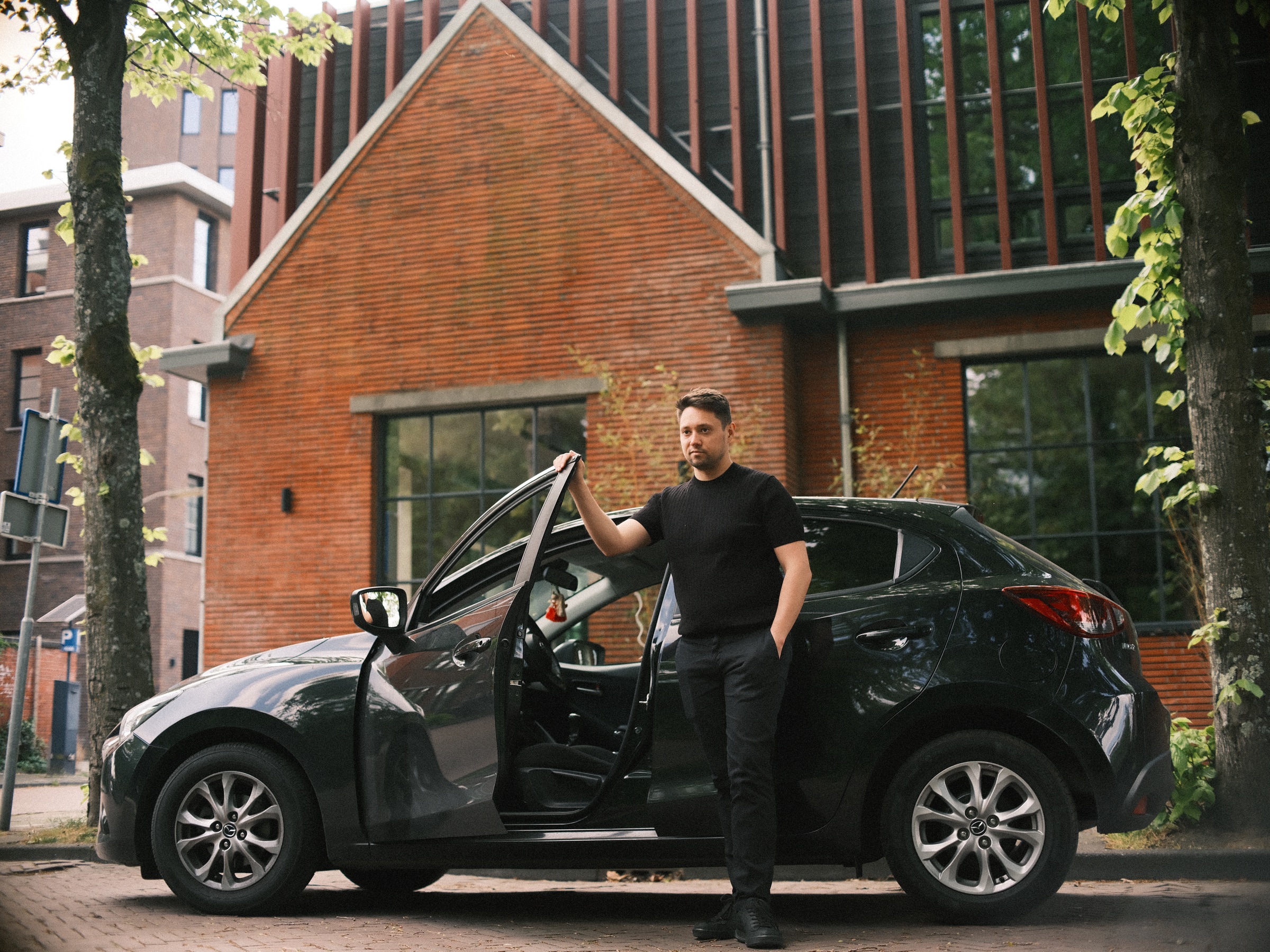 Man in het zwart staat bij het open portier van zijn donkere auto, geparkeerd voor een gebouw van rode baksteen met een spitse gevel