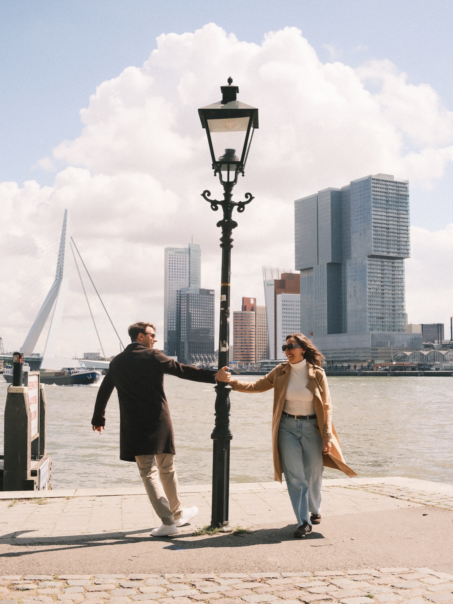 He swings around an old lamppost holding her hand on the quay, white clouds over the Erasmusbrug during their engagement photos