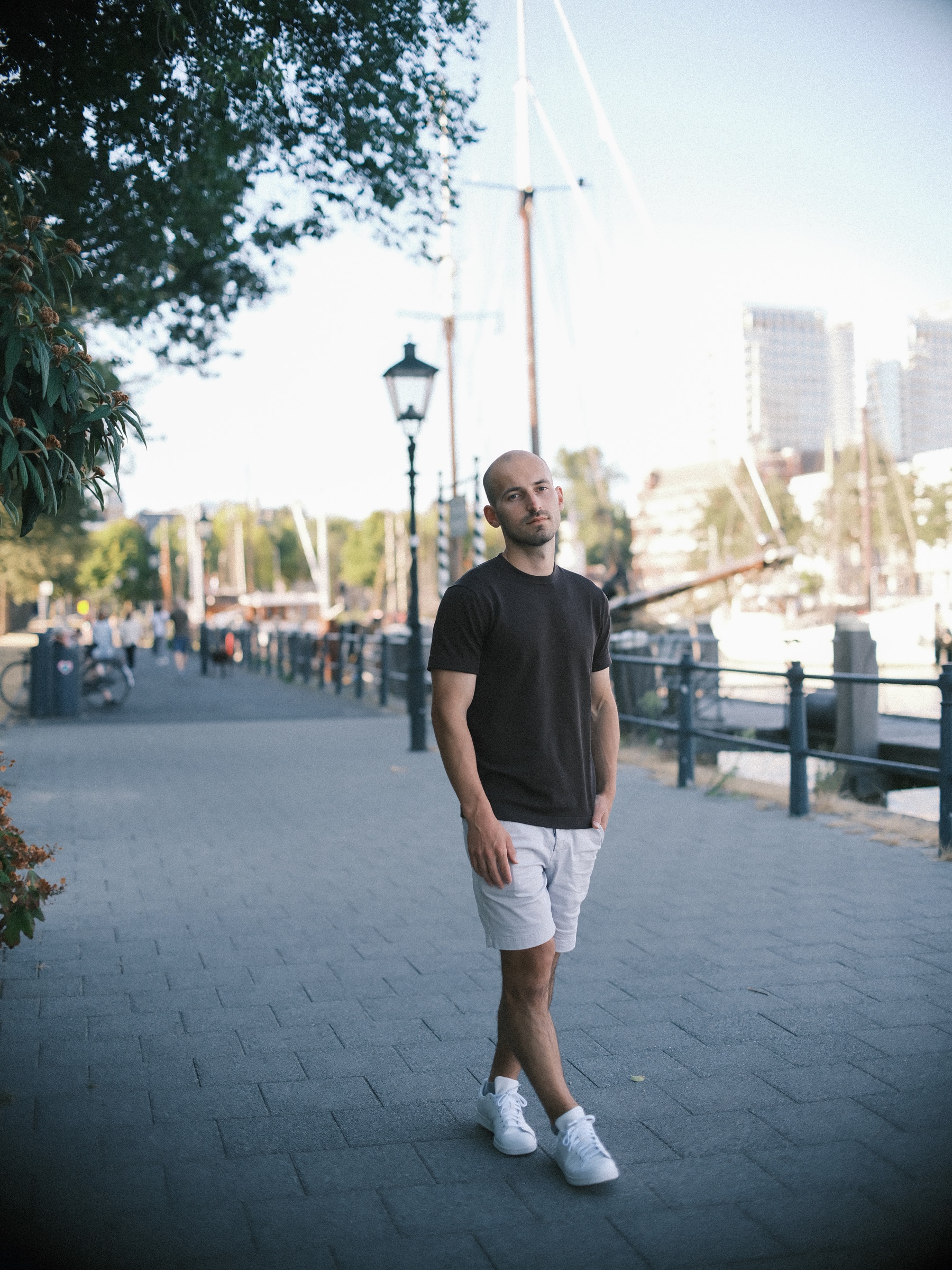 Man in a black T-shirt and light shorts on the Veerhaven quay on a summer evening, old lanterns, ship masts and towers behind him