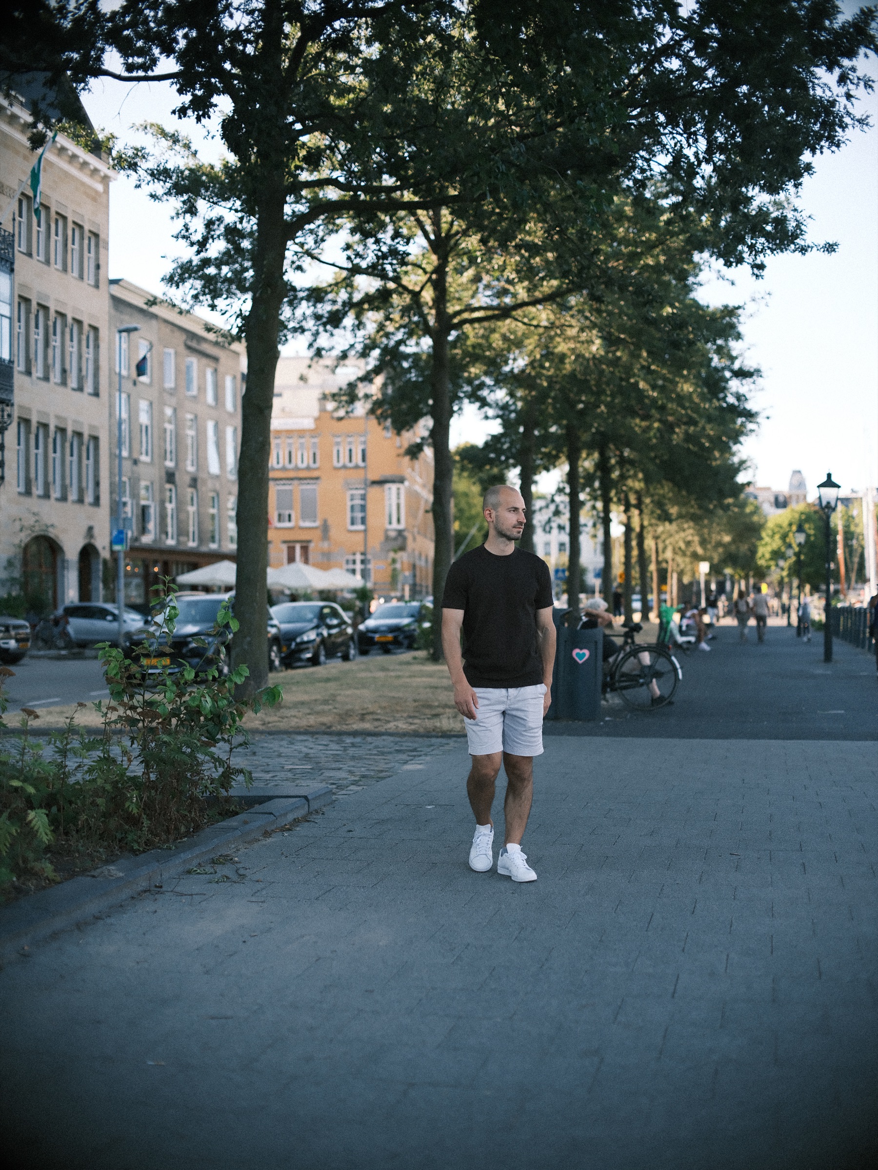 Hij loopt op de lens af door een zonnige straat met bomen en oude gevels, met warm avondlicht op de stoep