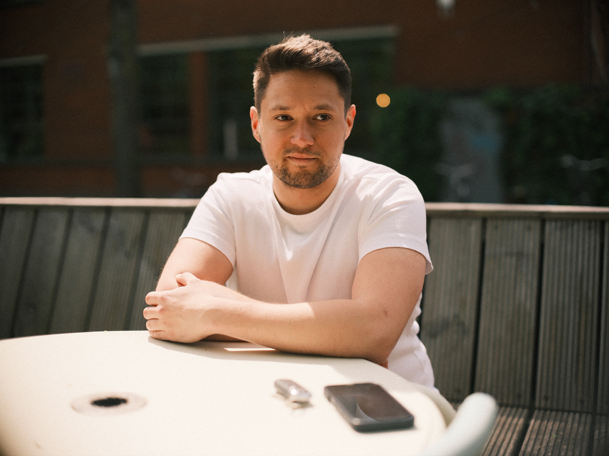 Man in een wit T-shirt aan een rond wit tafeltje op een zonnig terras in Haarlem, met zijn telefoon en sleutels naast zich, tijdens een solo fotoshoot