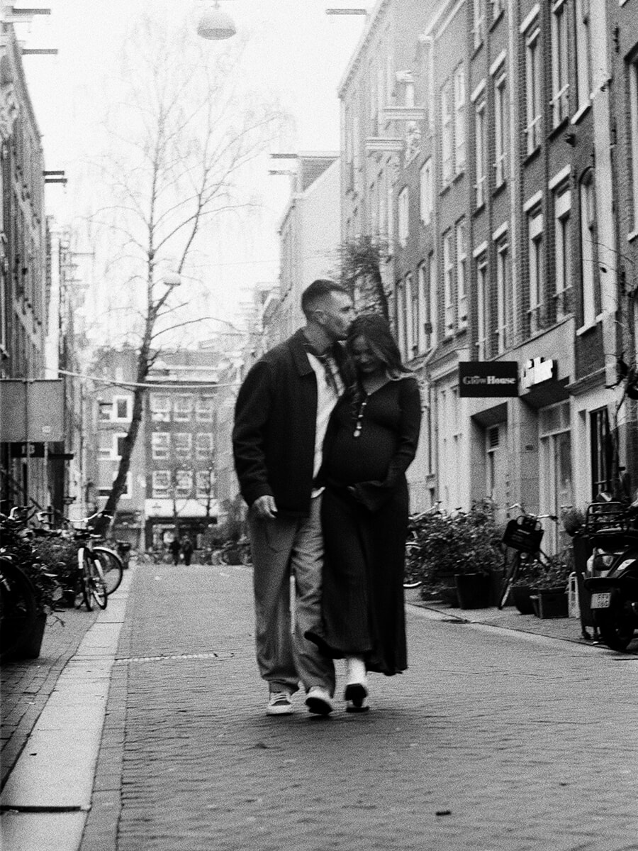 A man kisses his pregnant partner’s head as they walk along a narrow Amsterdam street during a maternity photoshoot on film