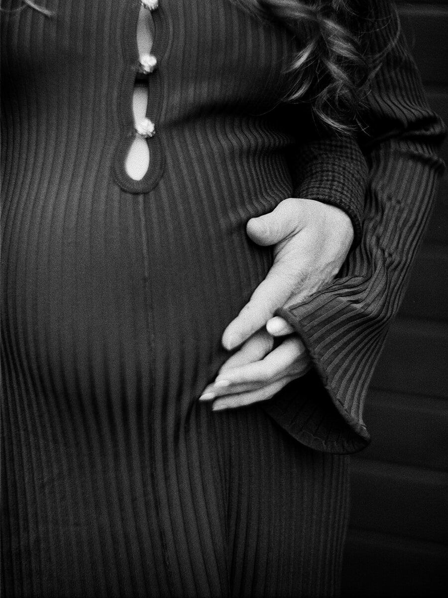 Close-up of hands resting together on a baby bump over a dark ribbed dress, captured on black-and-white film
