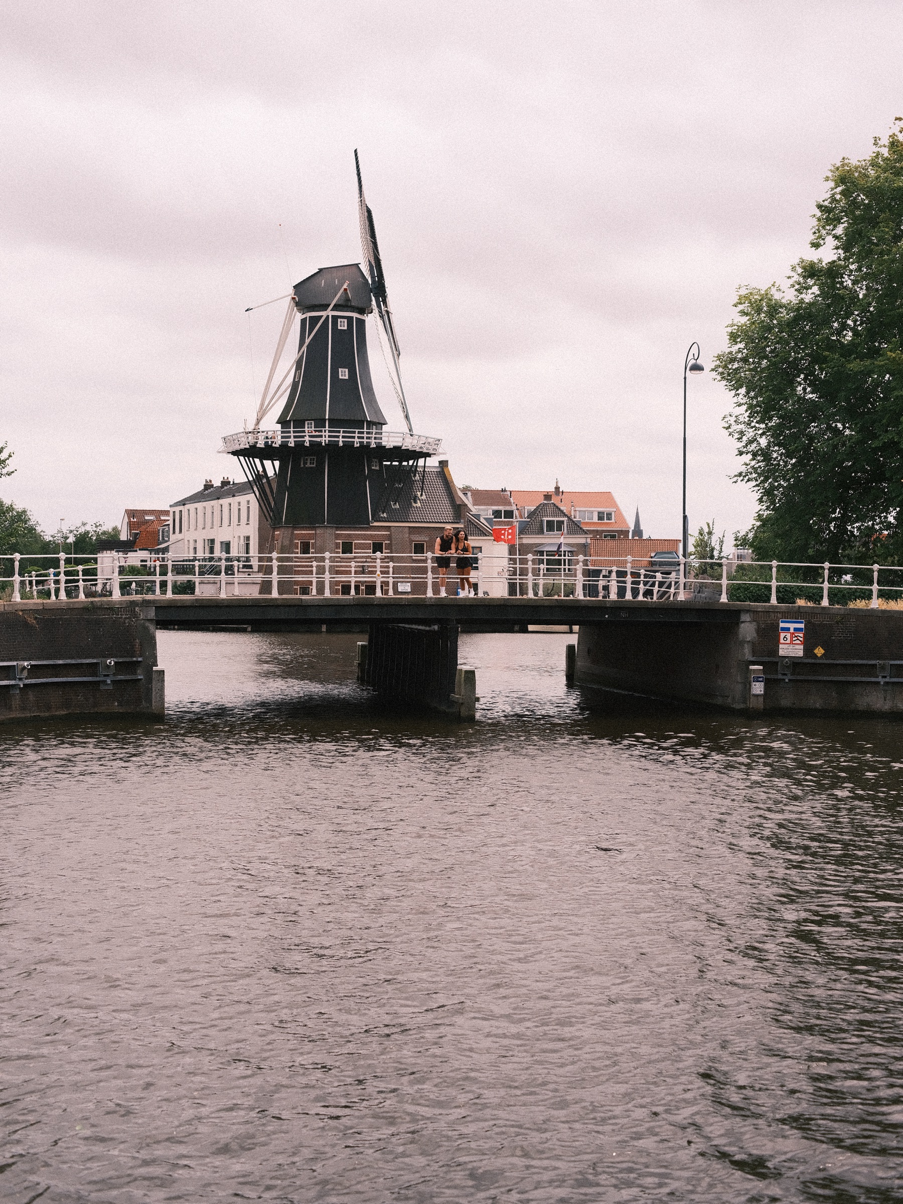Molen De Adriaan rijst op achter een lage brug over het Spaarne, de zwarte kap en wieken tegen een witte, bewolkte lucht
