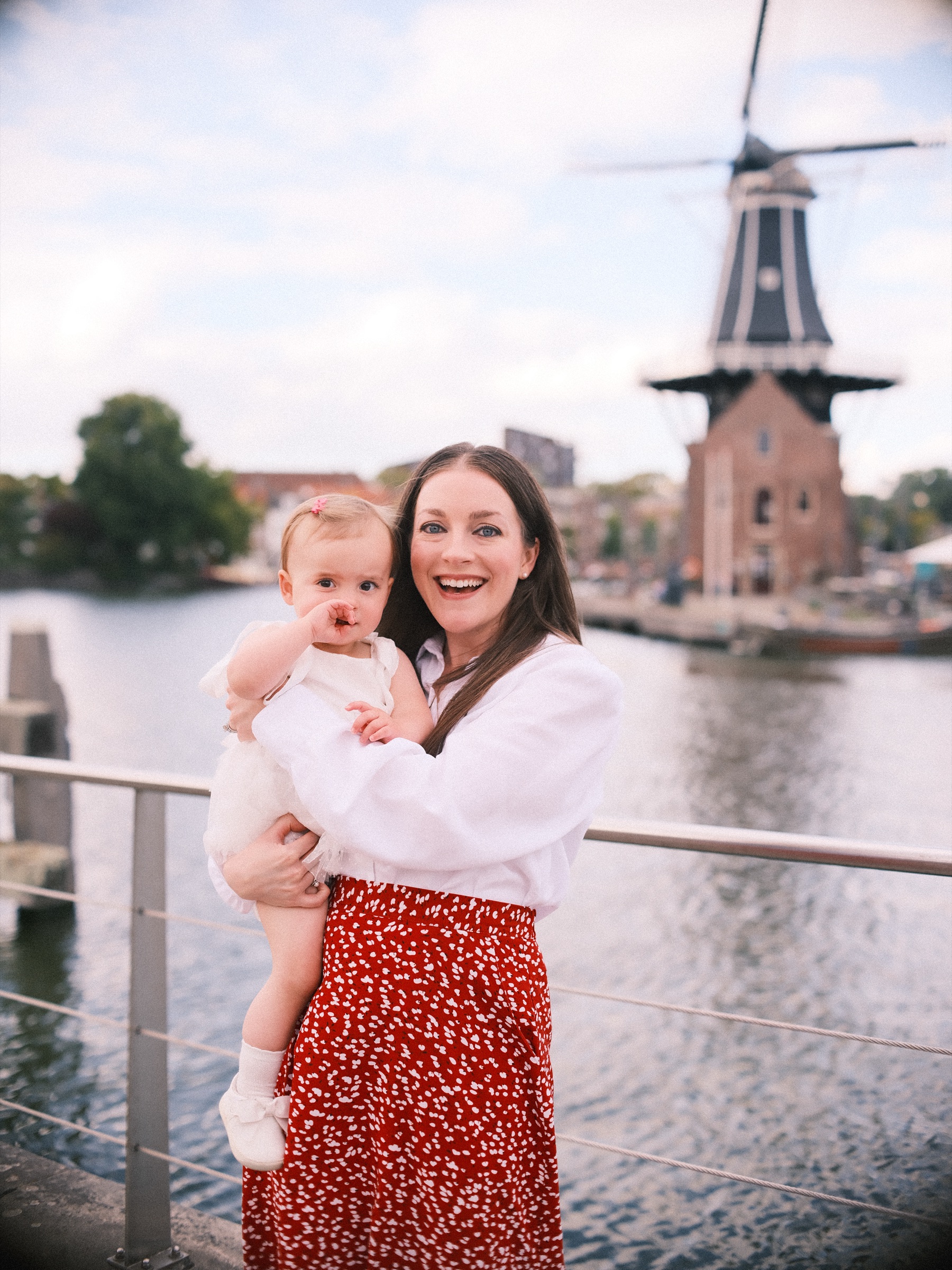 Moeder in een rode rok houdt haar dochtertje vast bij een reling aan het water, met molen De Adriaan onder witte wolken aan de overkant