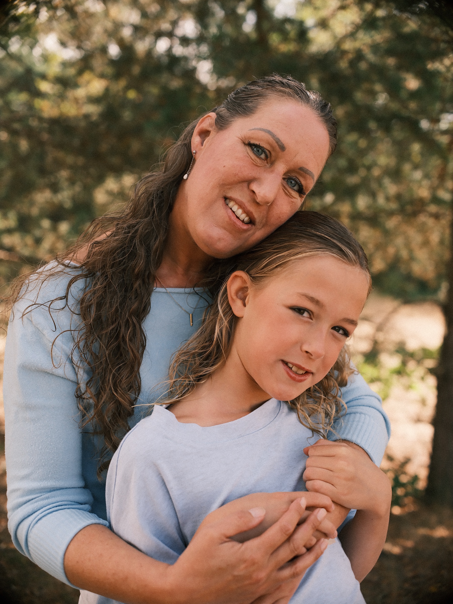 Mother in light blue hugging her daughter from behind, cheeks close together, both smiling among the pines