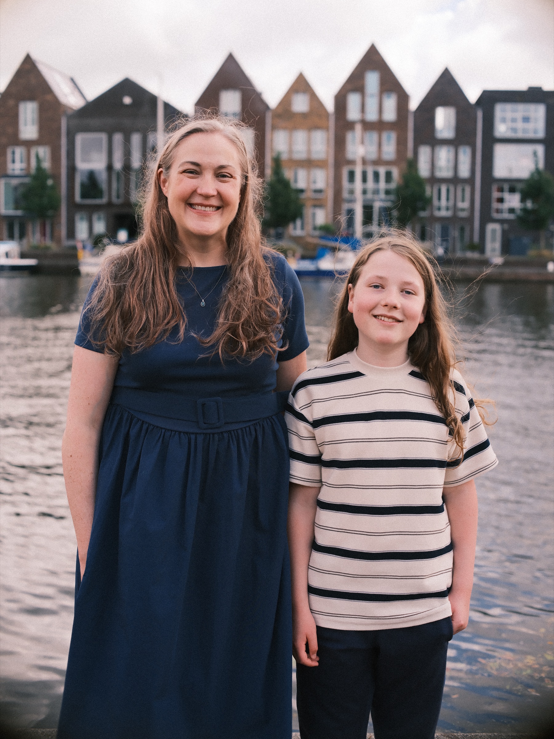 Moeder in een marineblauwe jurk en haar dochter in een gestreept T-shirt lachen aan het water, met de puntgevels van Haarlem erachter