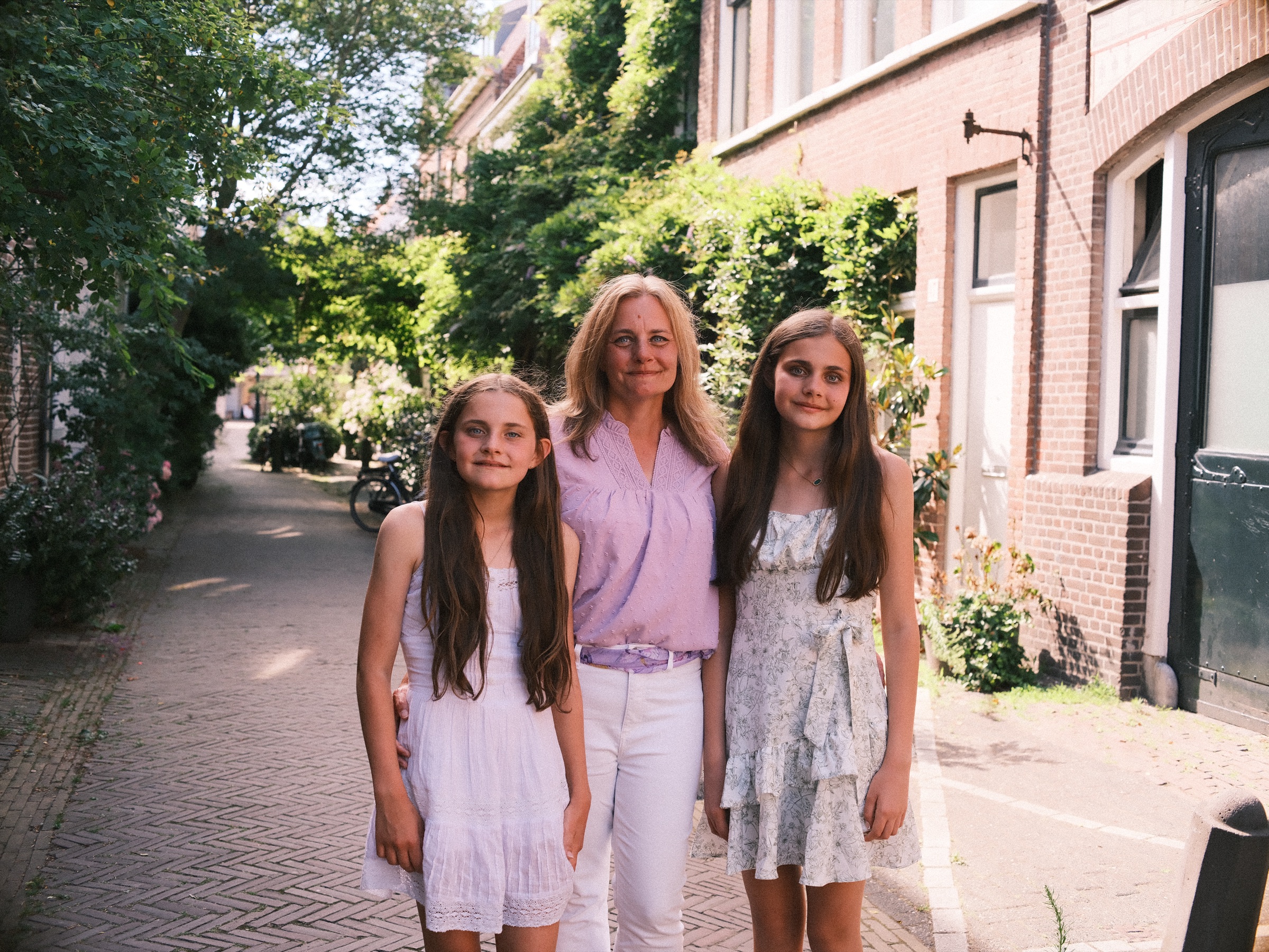 Moeder in lila tussen haar twee dochters in een groene straat met klinkers, klimplanten en hoge ramen langs de huizen in de zomerzon