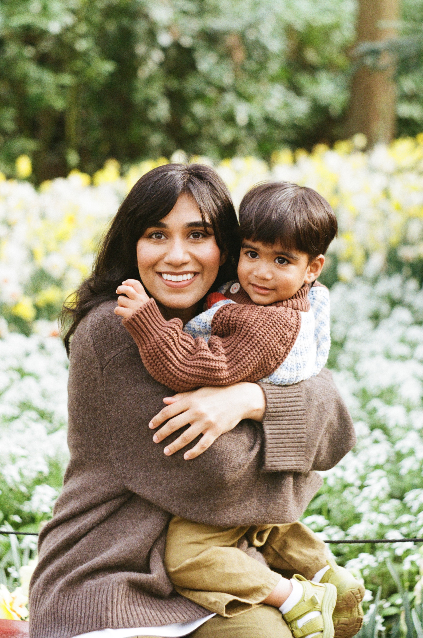 Smiling mother in a brown sweater hugging her toddler son close among white and yellow daffodils at Keukenhof