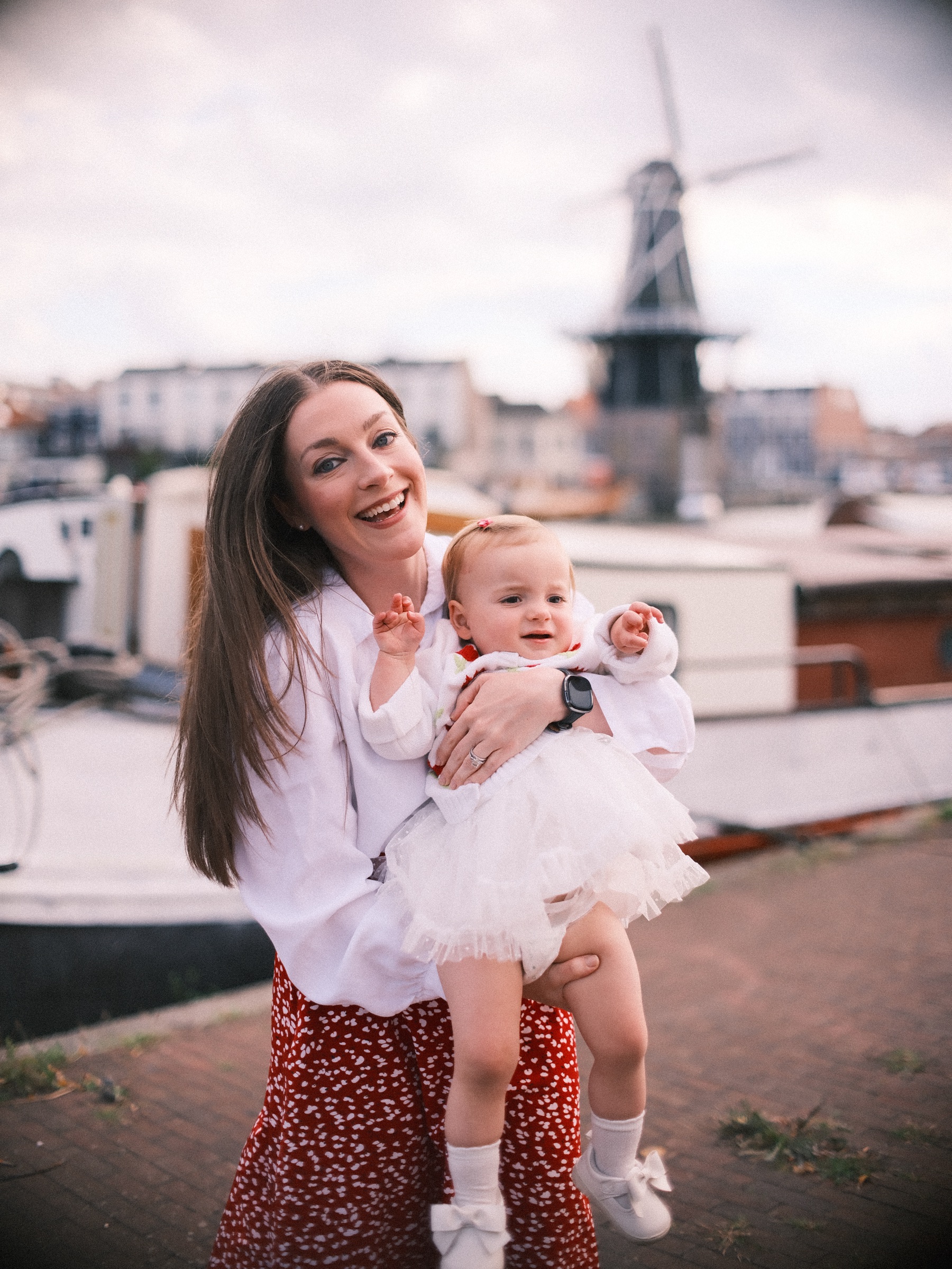 Moeder met lang donker haar lacht met haar dochtertje op haar heup, terwijl molen De Adriaan zacht op de achtergrond staat