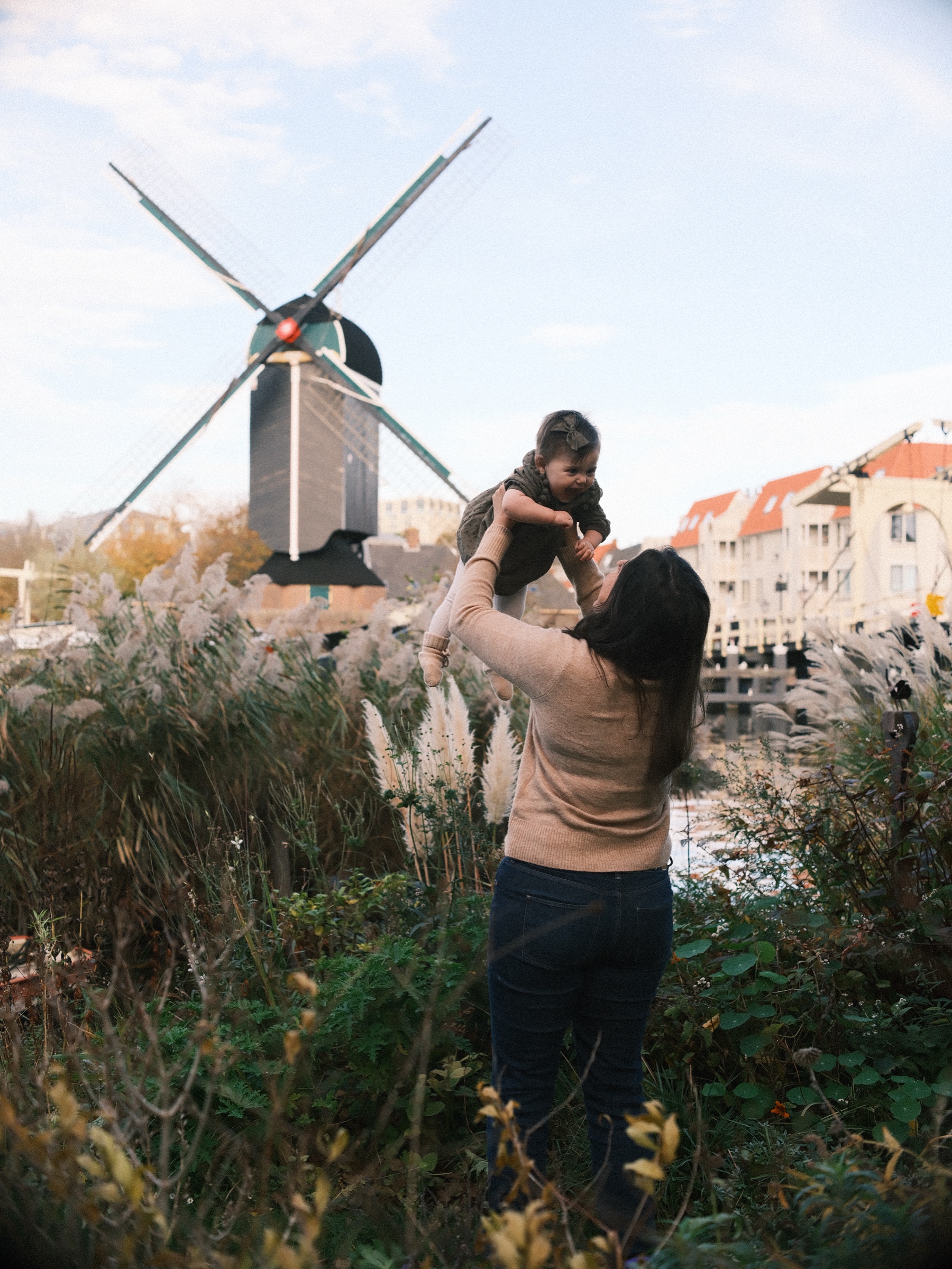 Mother holding her daughter up above her head by the water, the post mill and a row of white houses on the far bank