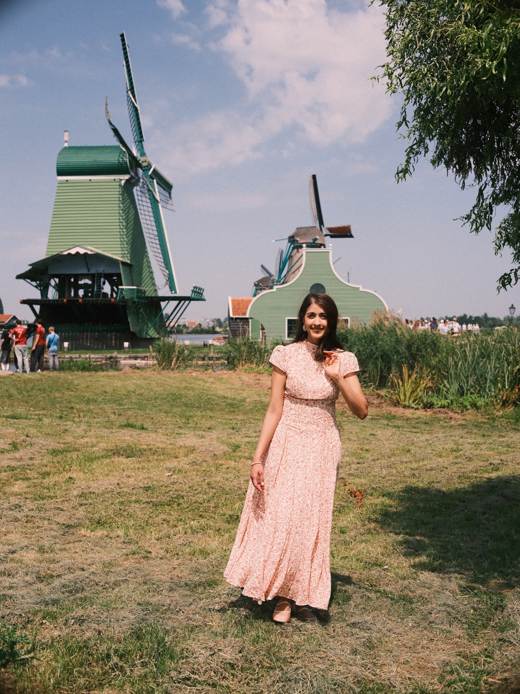 Mother in a long pink dress walking alone across a sunny meadow, the green windmill of the Zaanse Schans and a blue sky behind her