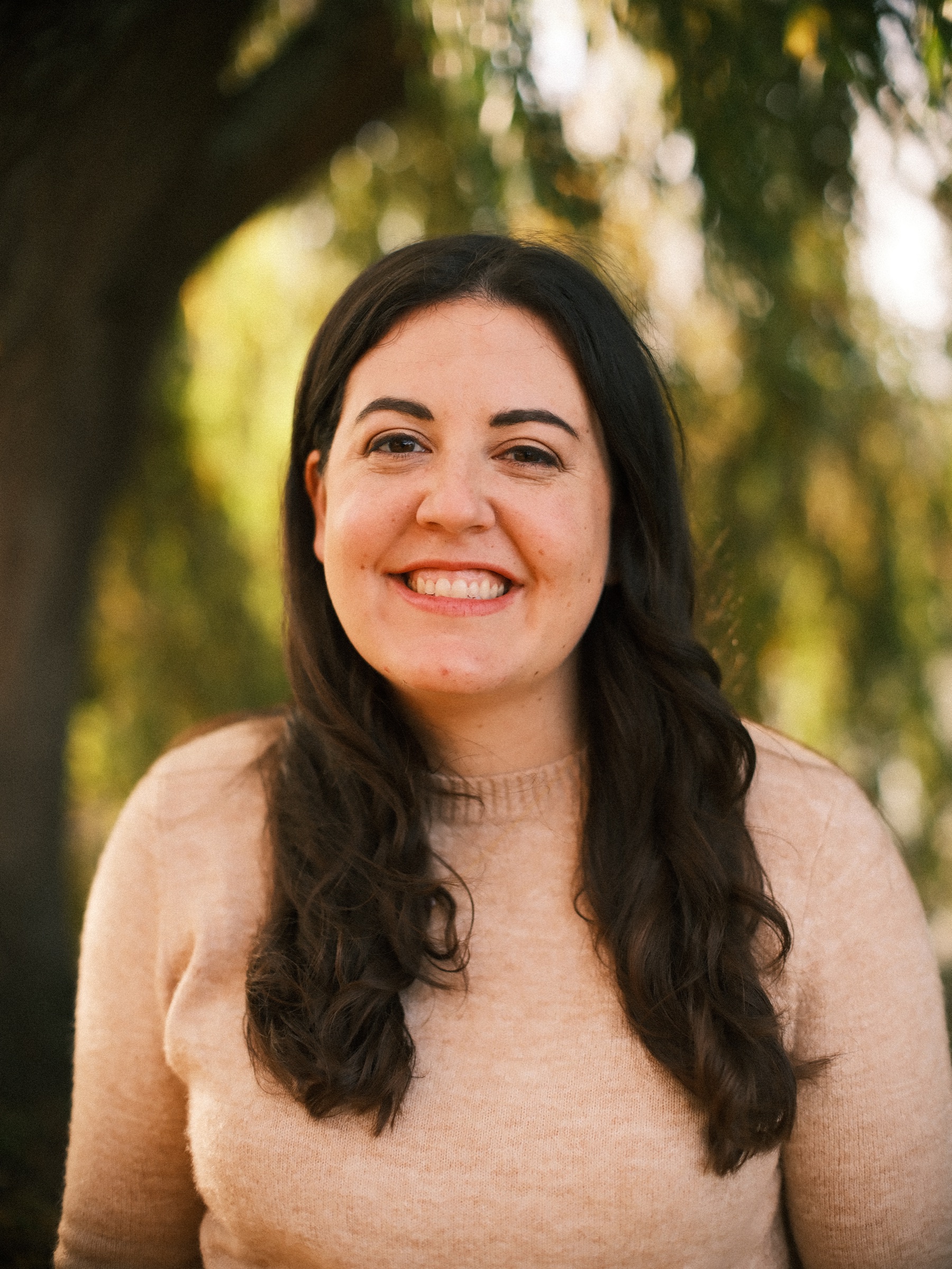Close portrait of a smiling woman with long dark hair in a peach sweater, the yellowing branches of a willow glowing behind her