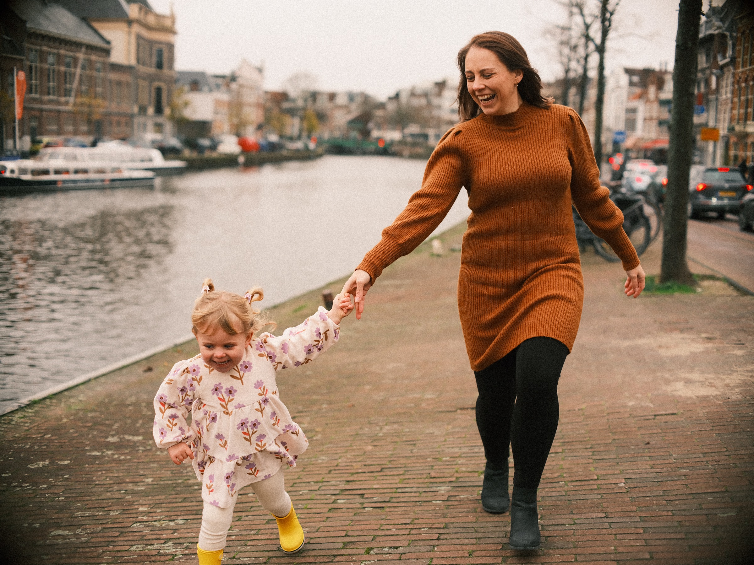 Moeder rent over de kade van het Spaarne met haar lachende dochtertje aan de hand, met boten en gevels erachter