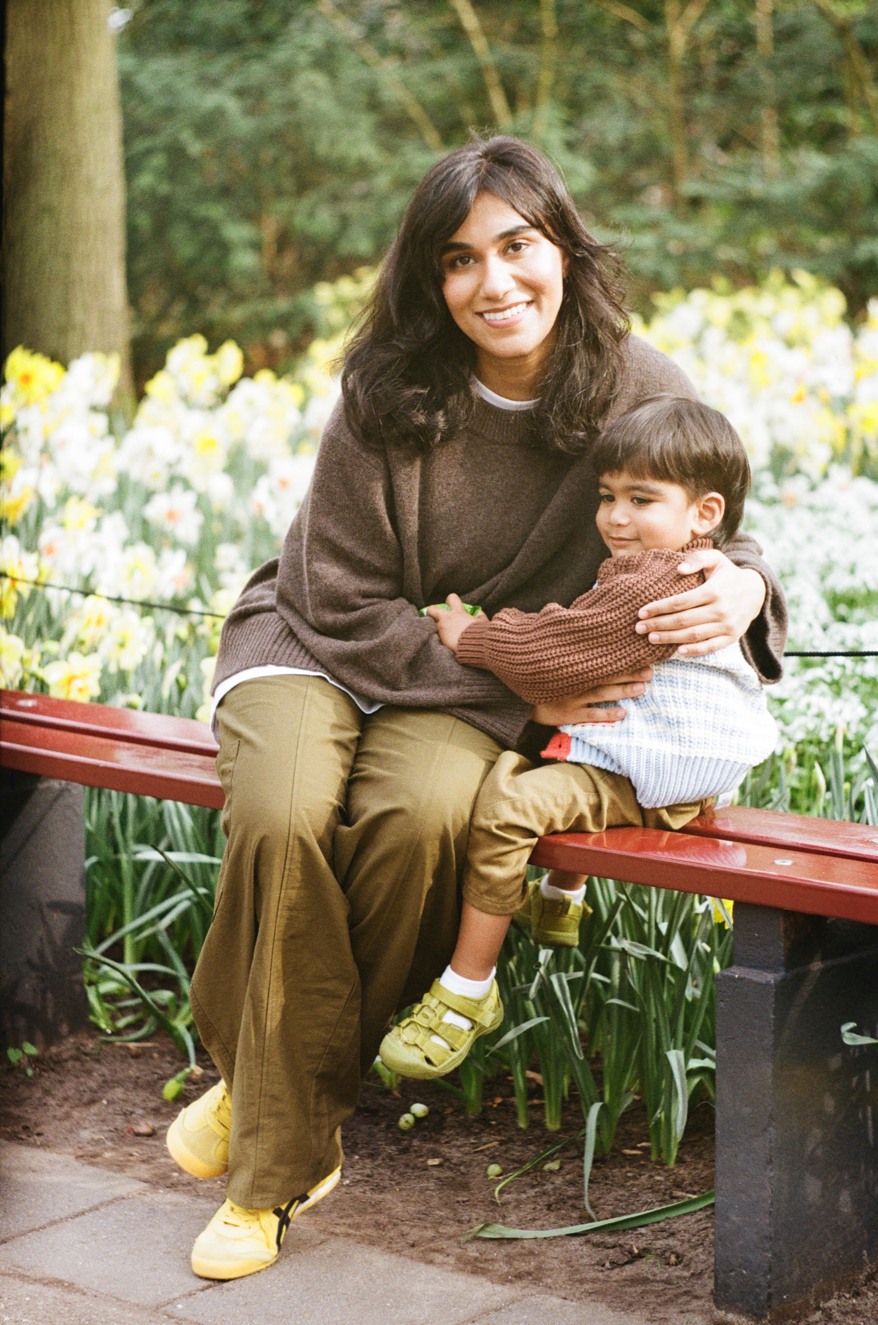 Mother on a red bench with her little boy on her lap, both smiling, her yellow shoes bright against the daffodil beds