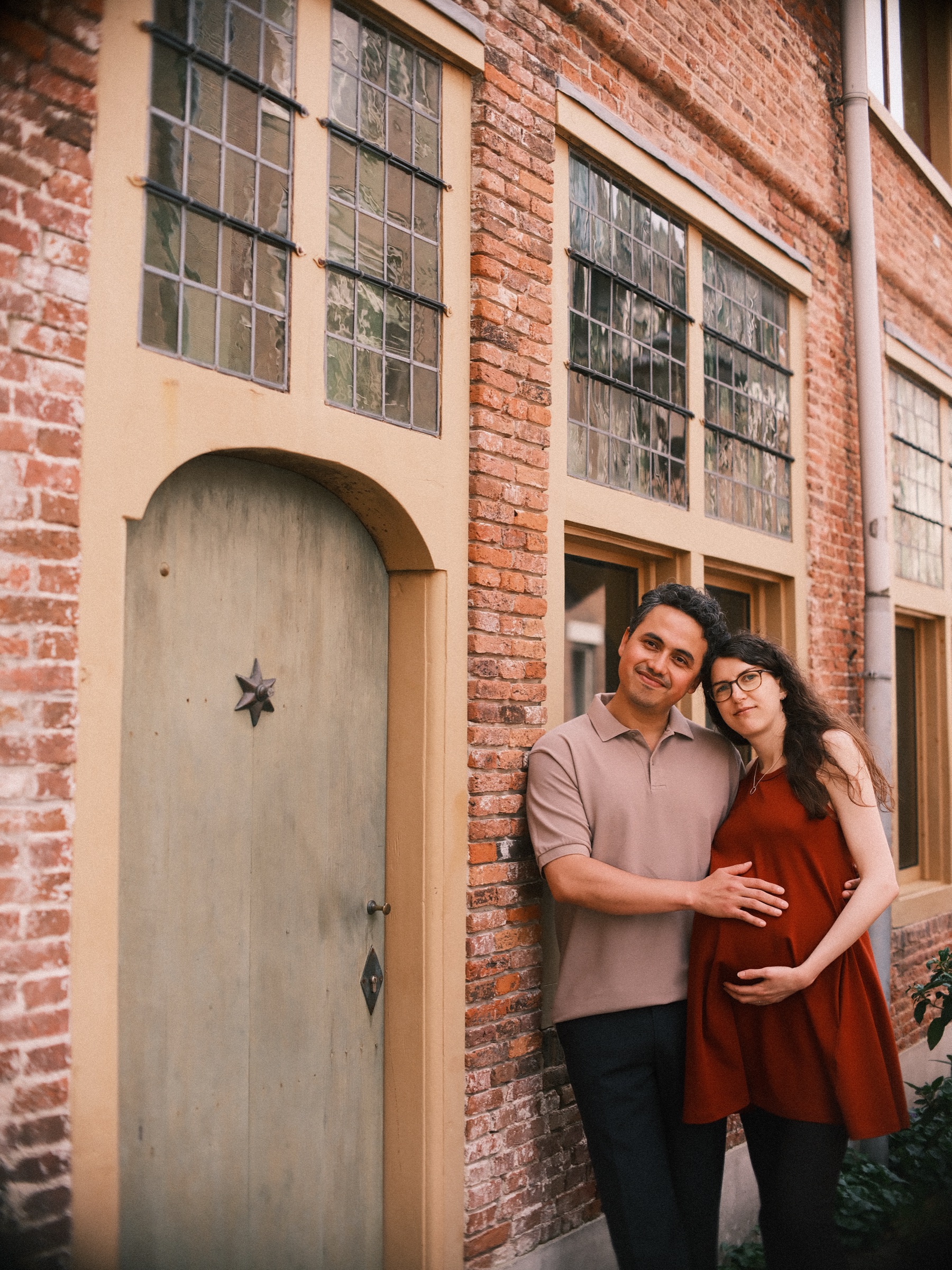 Laughing mother-to-be leaning into her partner by a pale arched door with a small star on it, his hand on her bump