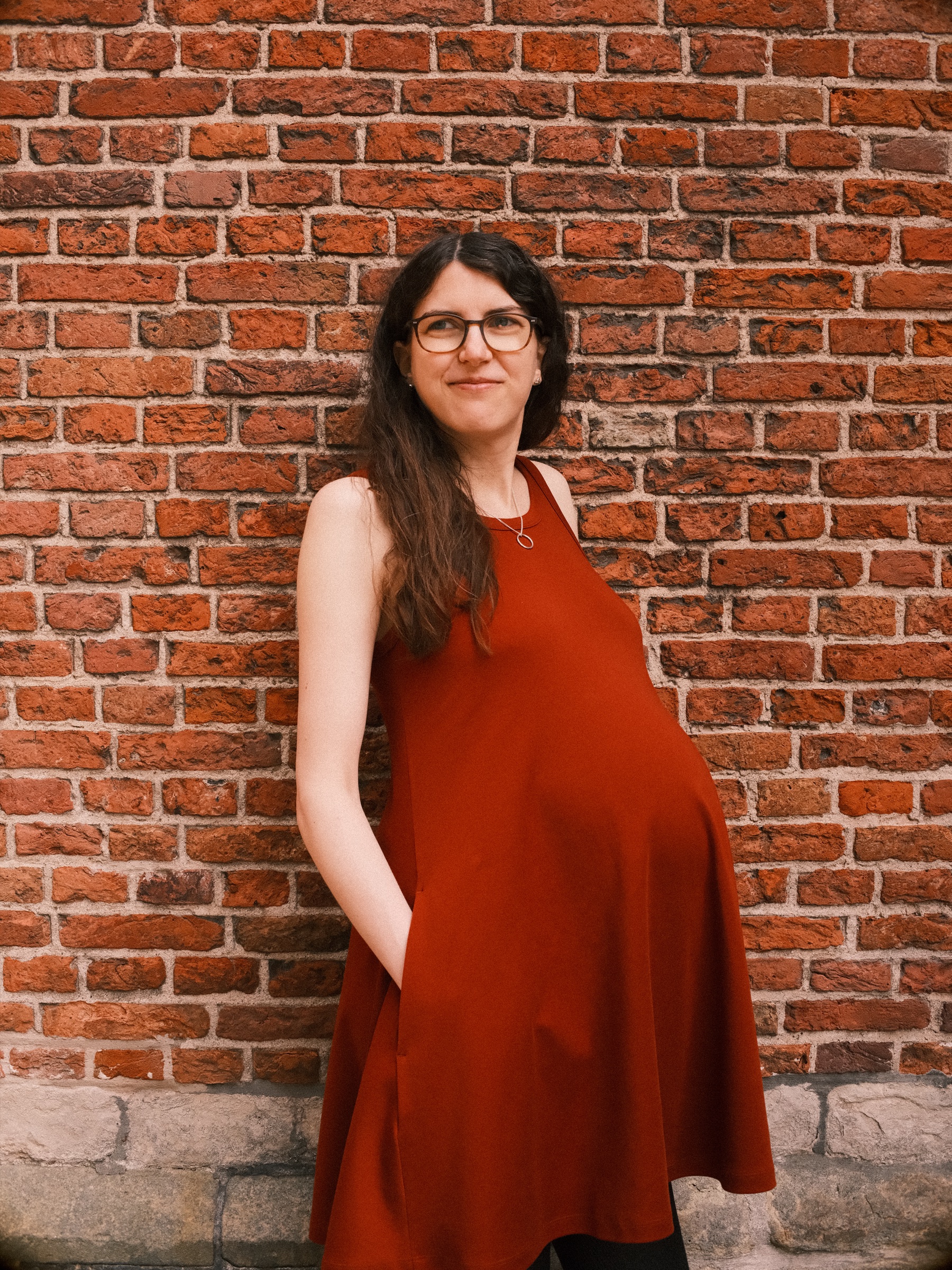 Mother-to-be in a sleeveless rust-red dress smiling against old red brick, her long dark hair over one shoulder and her baby bump in profile