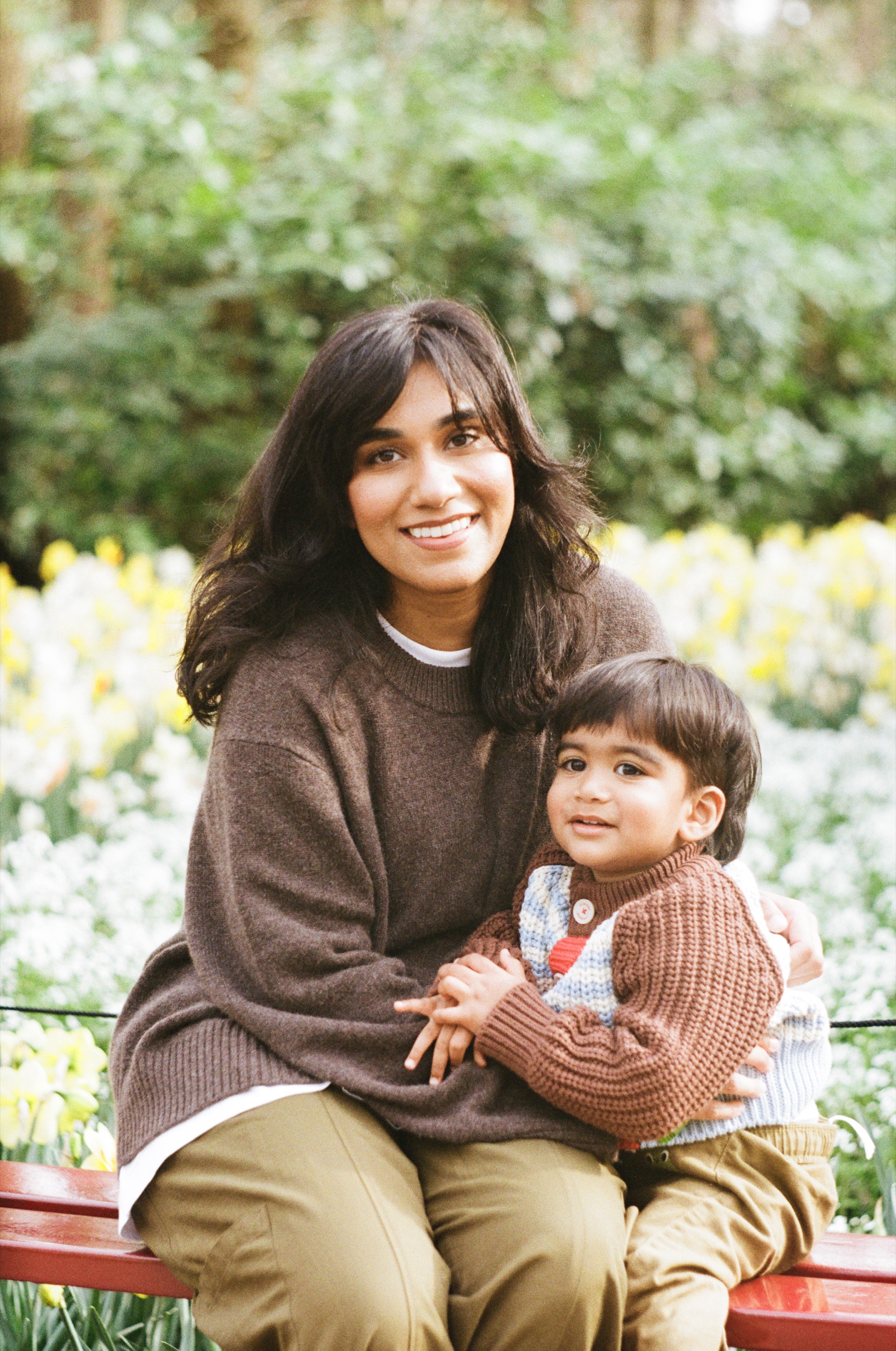Smiling mother in a brown sweater with her toddler on her lap on a red bench, daffodils behind them, a film photoshoot on Kodak Gold 200