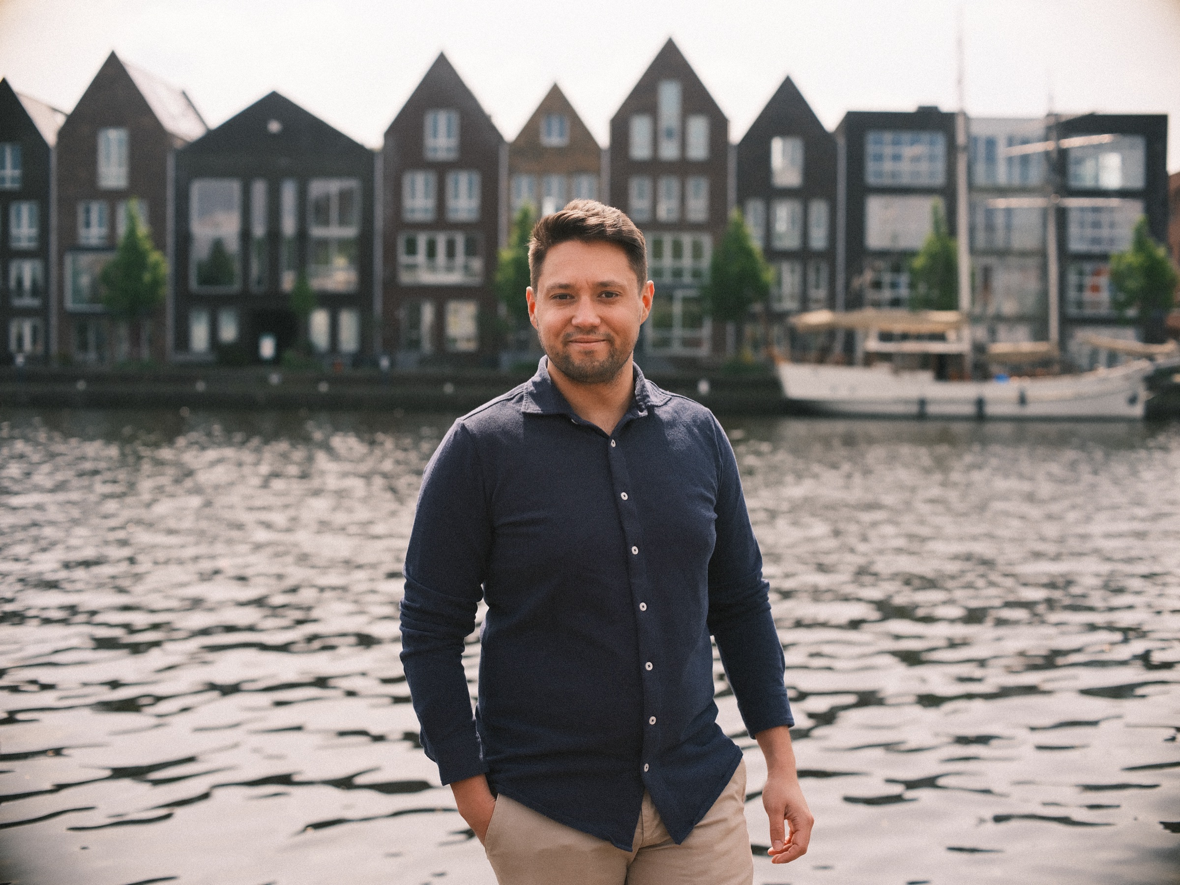 Man in donkerblauw overhemd en lichte broek aan het Spaarne in Haarlem, met nieuwe huizen met puntgevels aan de overkant