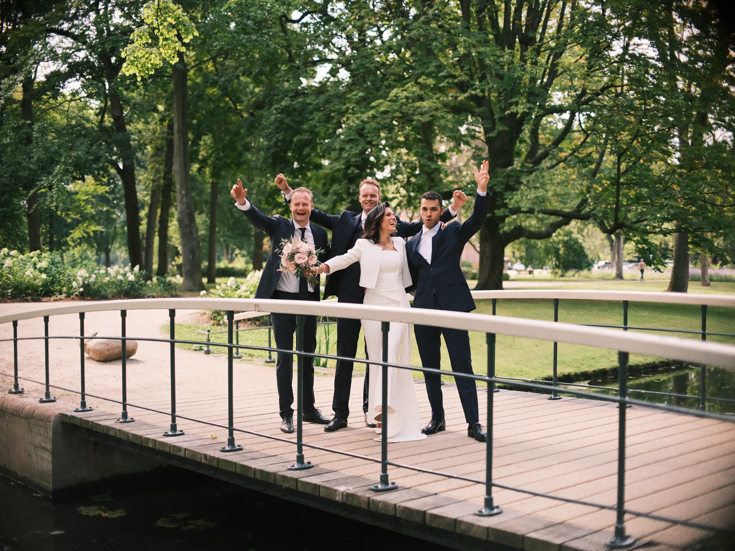Bruidspaar juicht met twee familieleden op een wit bruggetje in Het Park, met de armen omhoog onder hoge bomen
