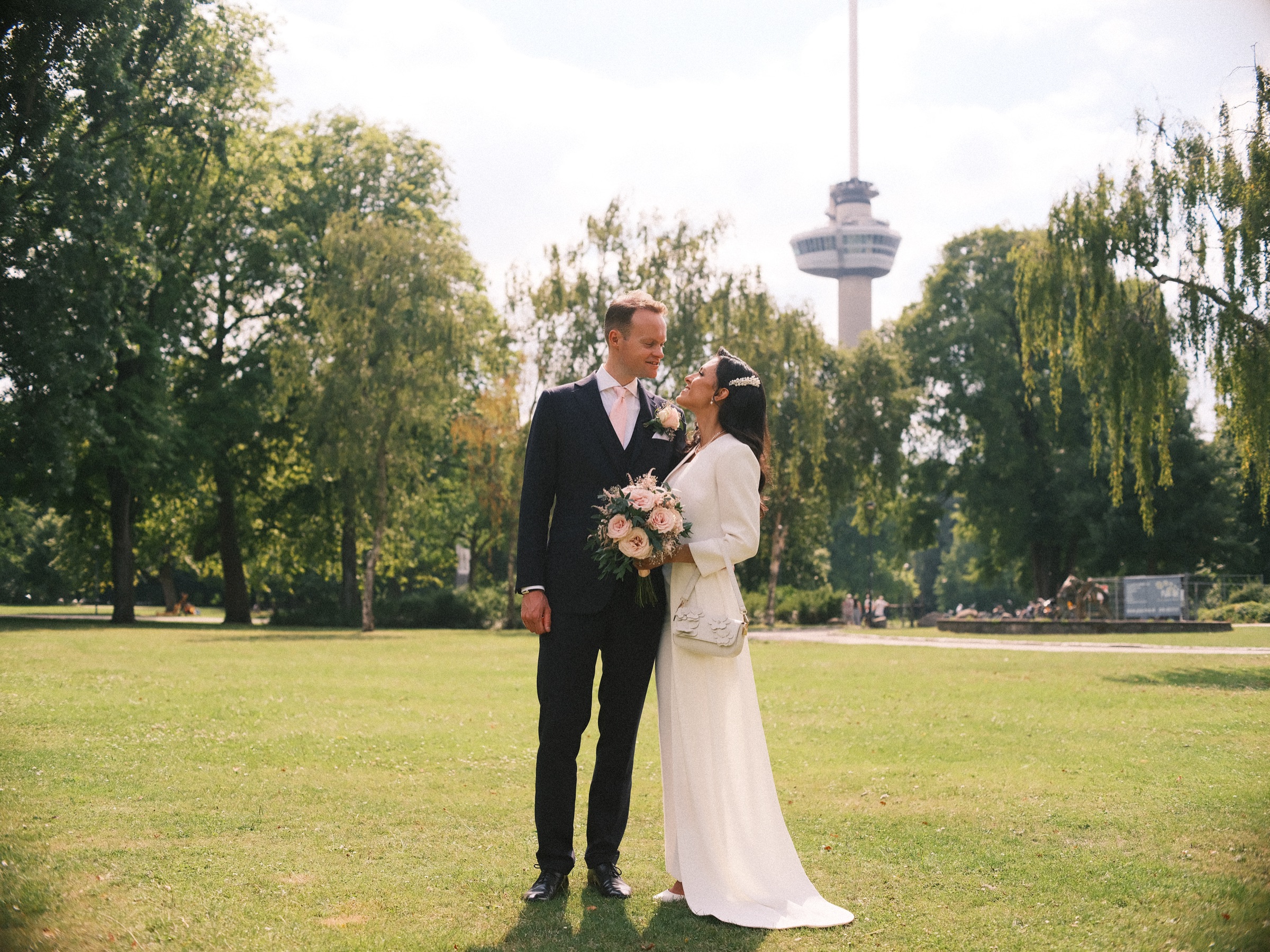 Bruid en bruidegom op een groot grasveld met de Euromast hoog boven de bomen, een trouwfoto in Het Park in de zomer