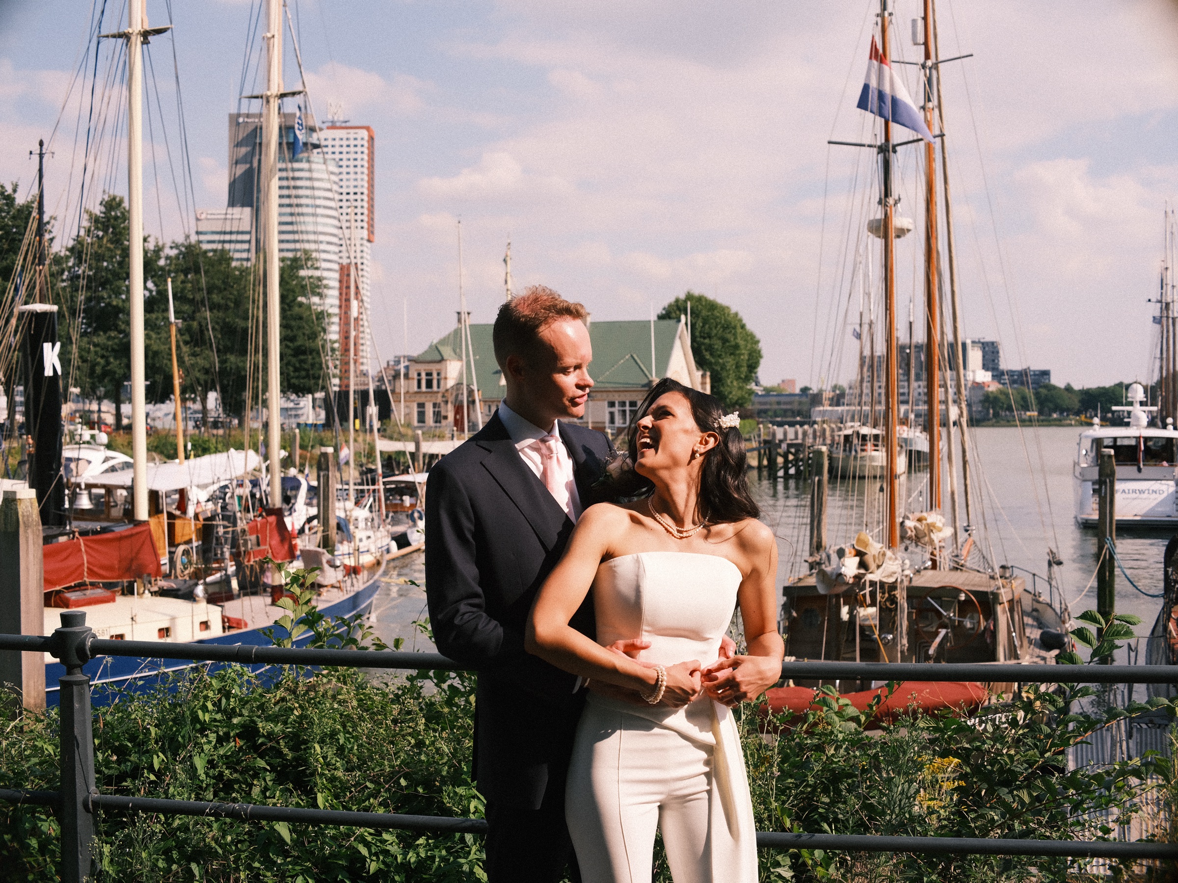 Bruid en bruidegom lachen voor hoge zeilschepen met masten en een Nederlandse vlag in de Veerhaven, in de zon