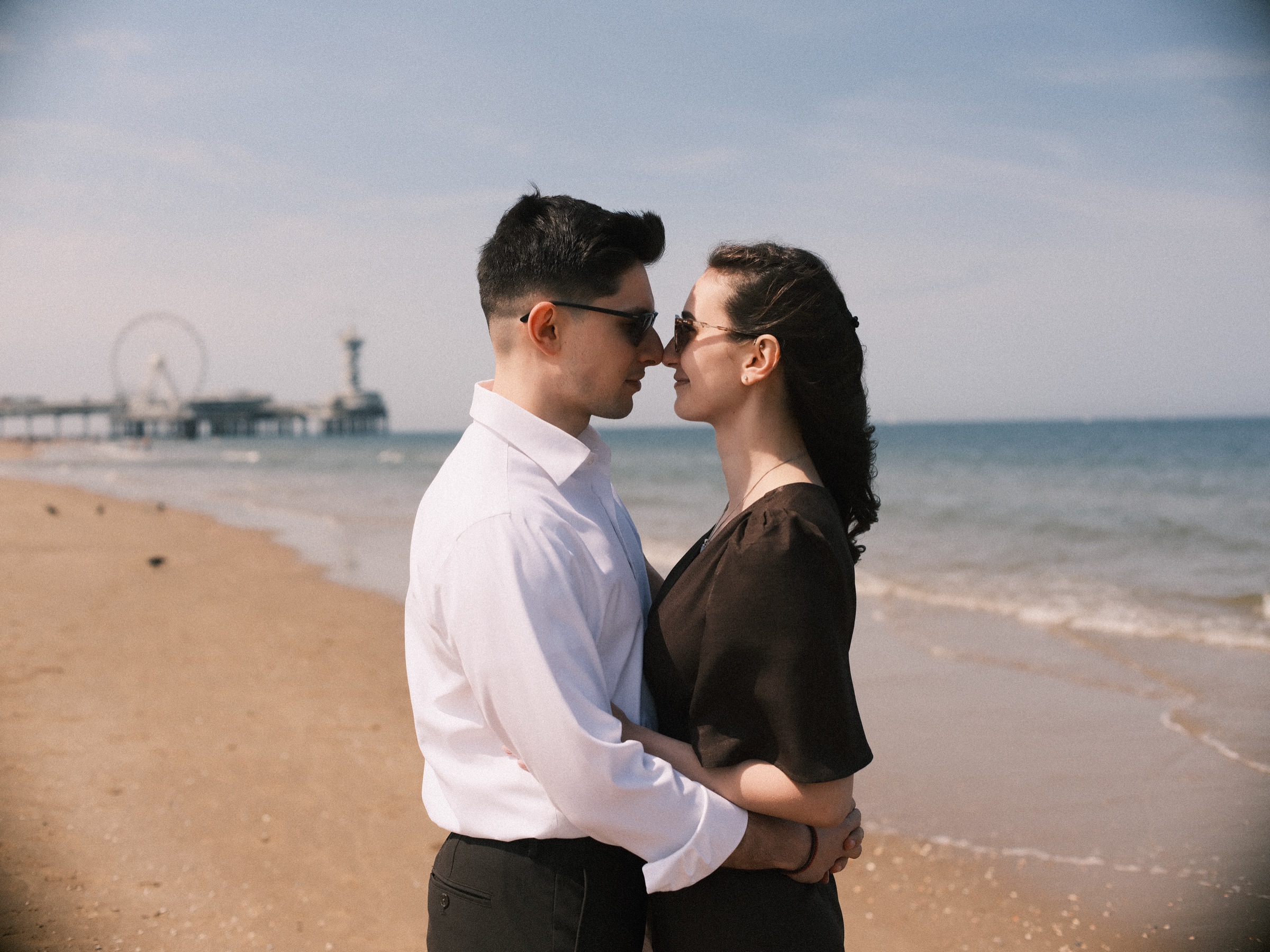 Couple standing nose to nose in sunglasses on Scheveningen beach, the Pier and its Ferris wheel soft in the distance