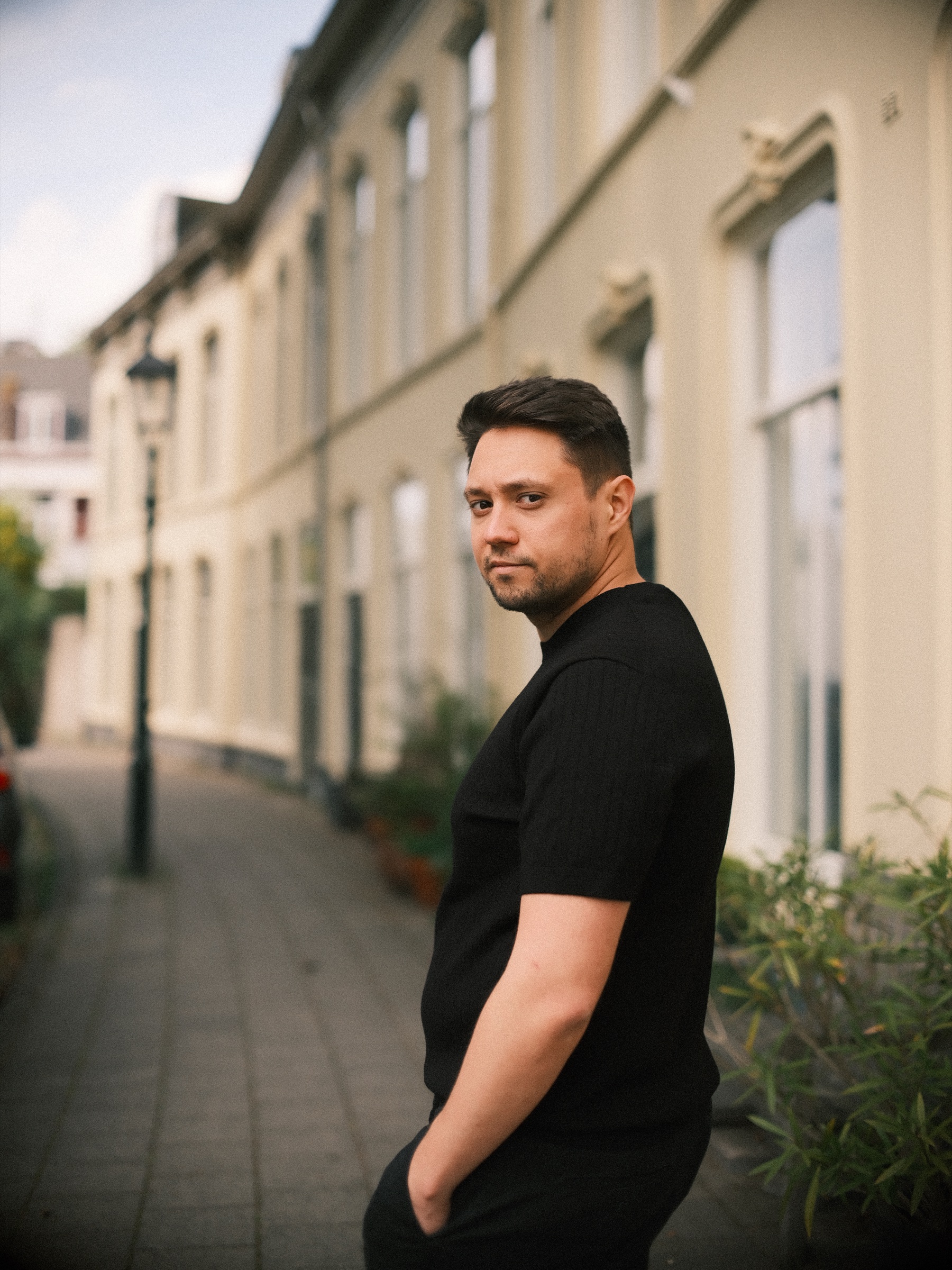 Man in zwart T-shirt kijkt over zijn schouder in een stille straat met crèmekleurige huizen, een oude lantaarn en planten