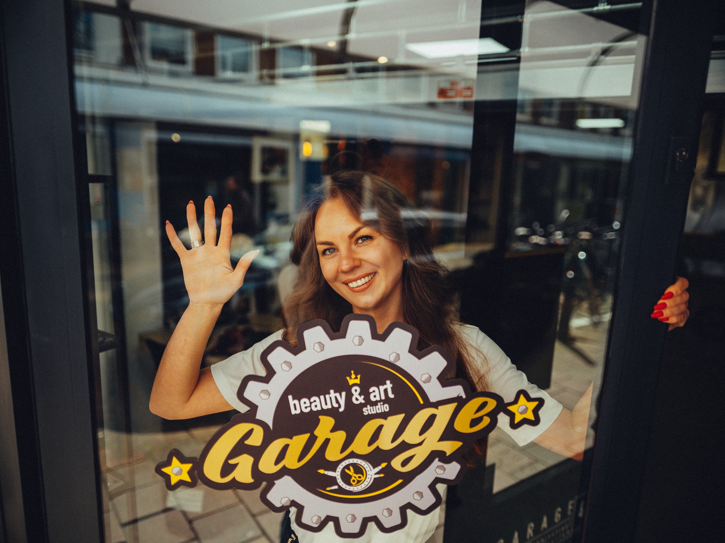 Eigenaar zwaait achter het raam van haar beautystudio, met het logo van Garage en een weerspiegelde straat in het glas