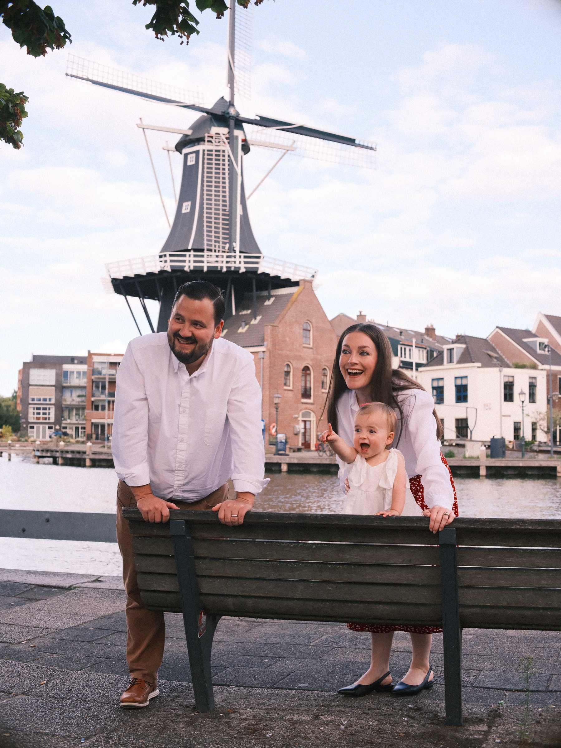 Vader in een wit overhemd leunt op een bank terwijl de moeder lacht met hun baby, met molen De Adriaan en oude huizen erachter