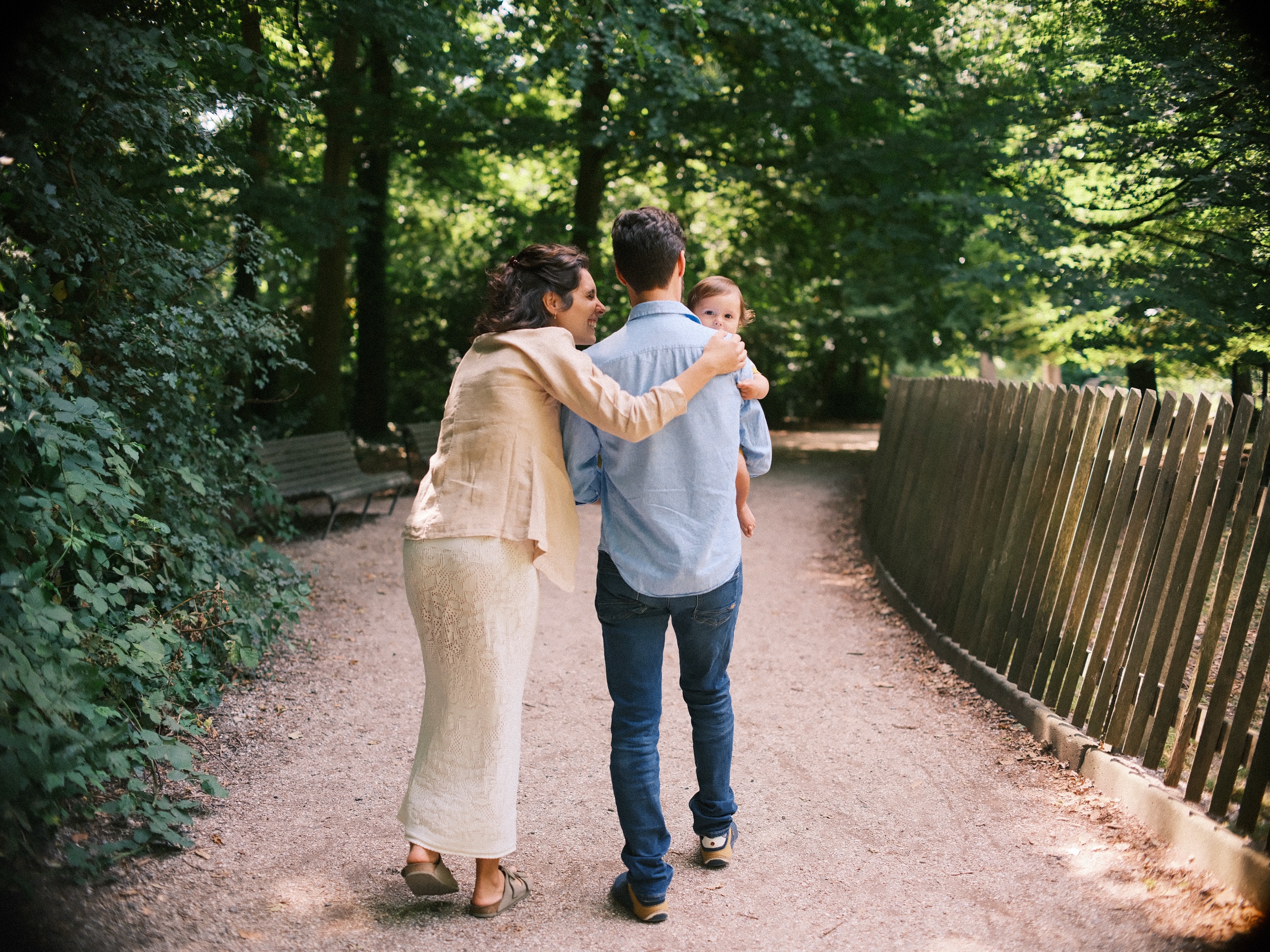 Ouders lopen over een grindpad langs een houten hek, de vader met de baby op de arm en de moeder met een hand op zijn rug