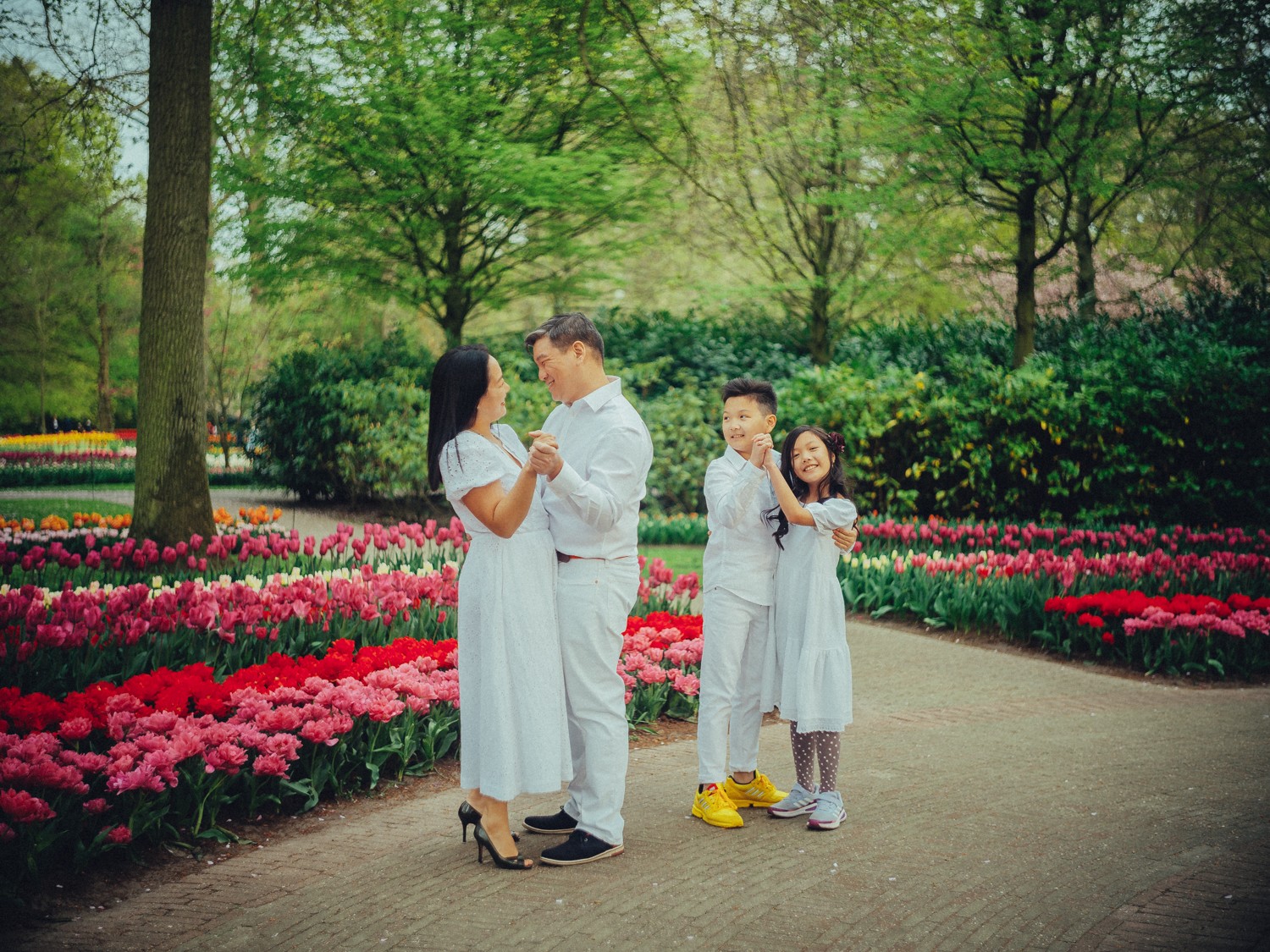 Ouders dansen op een tuinpad terwijl hun zoon met gele sneakers zijn zusje laat draaien, tussen perken met roze en rode tulpen