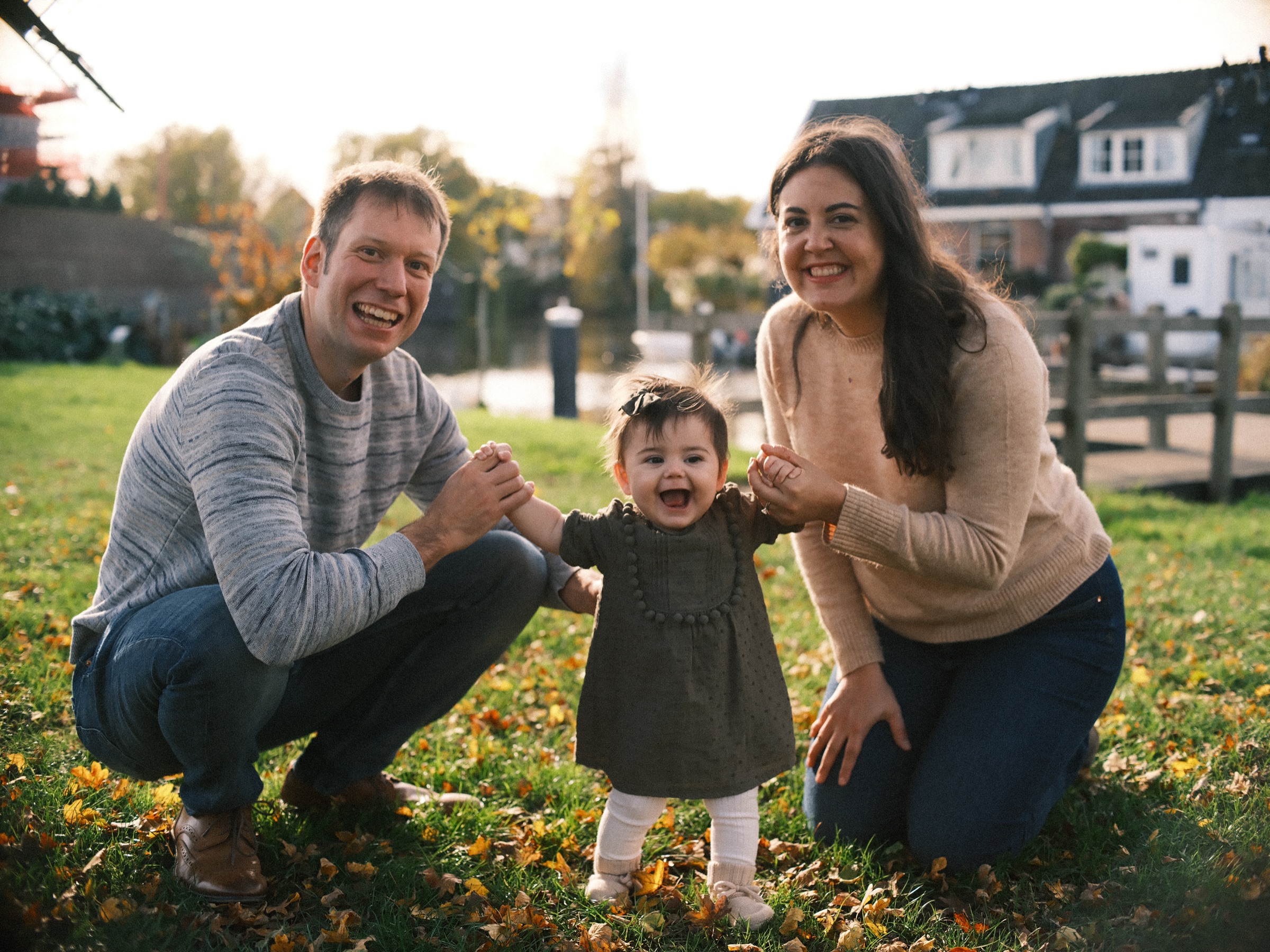 Parents crouching in the grass in Leiden while their baby stands between them, holding their hands on a bright autumn day