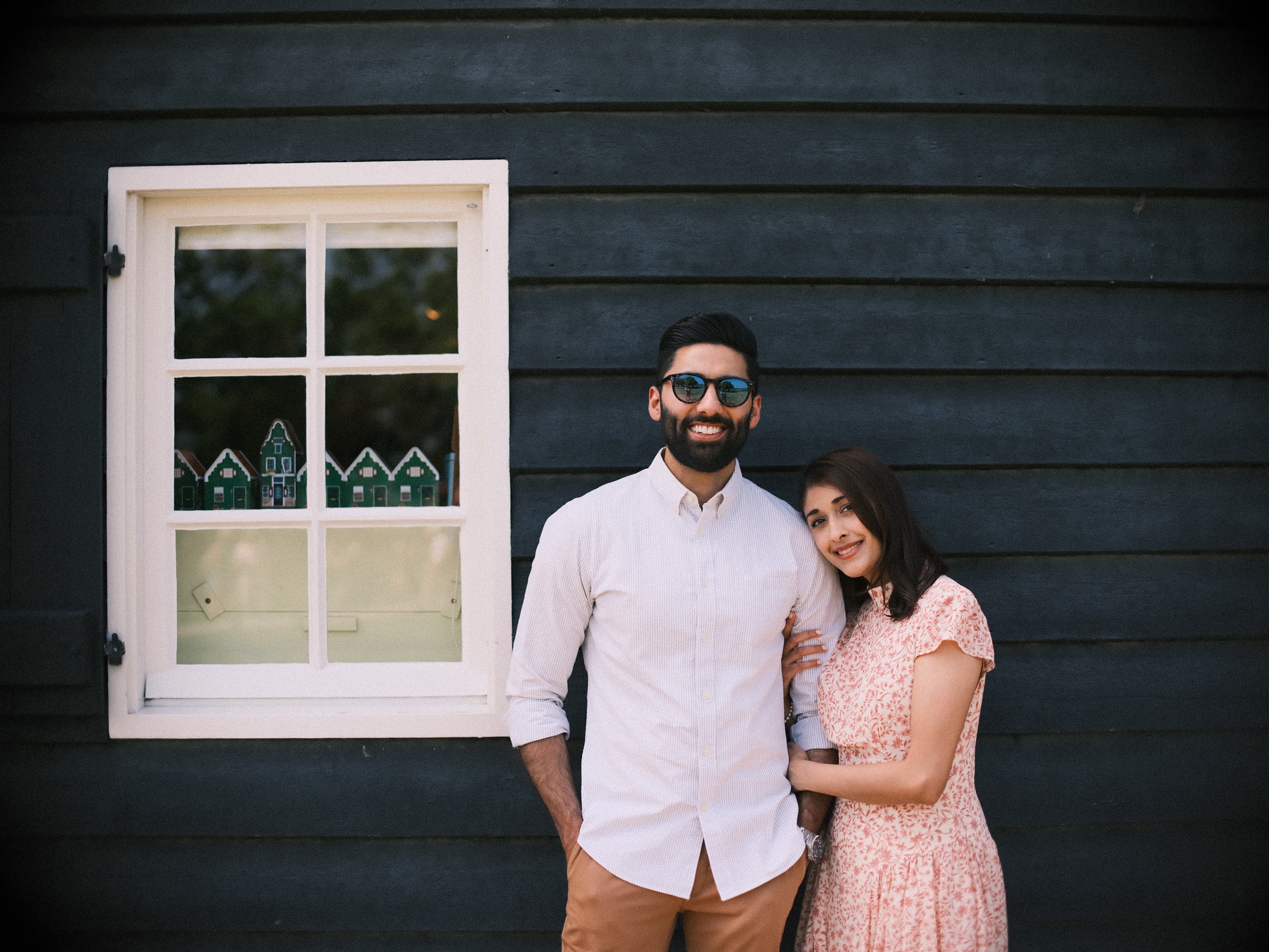 Couple standing close against a dark wooden wall beside a white window full of tiny green houses, both smiling in the sun